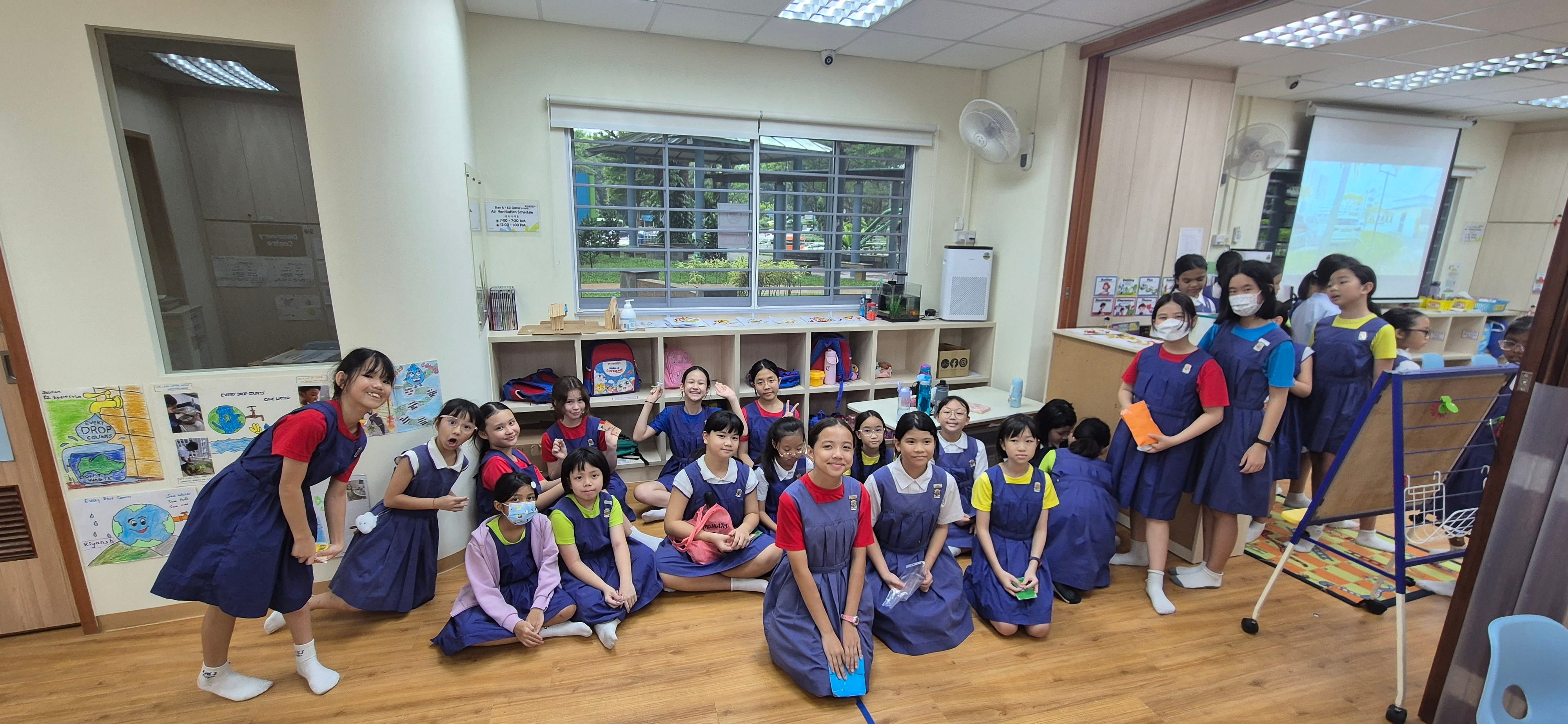 A group of young schoolgirls in blue uniforms sitting and standing on a classroom floor, smiling at the camera.