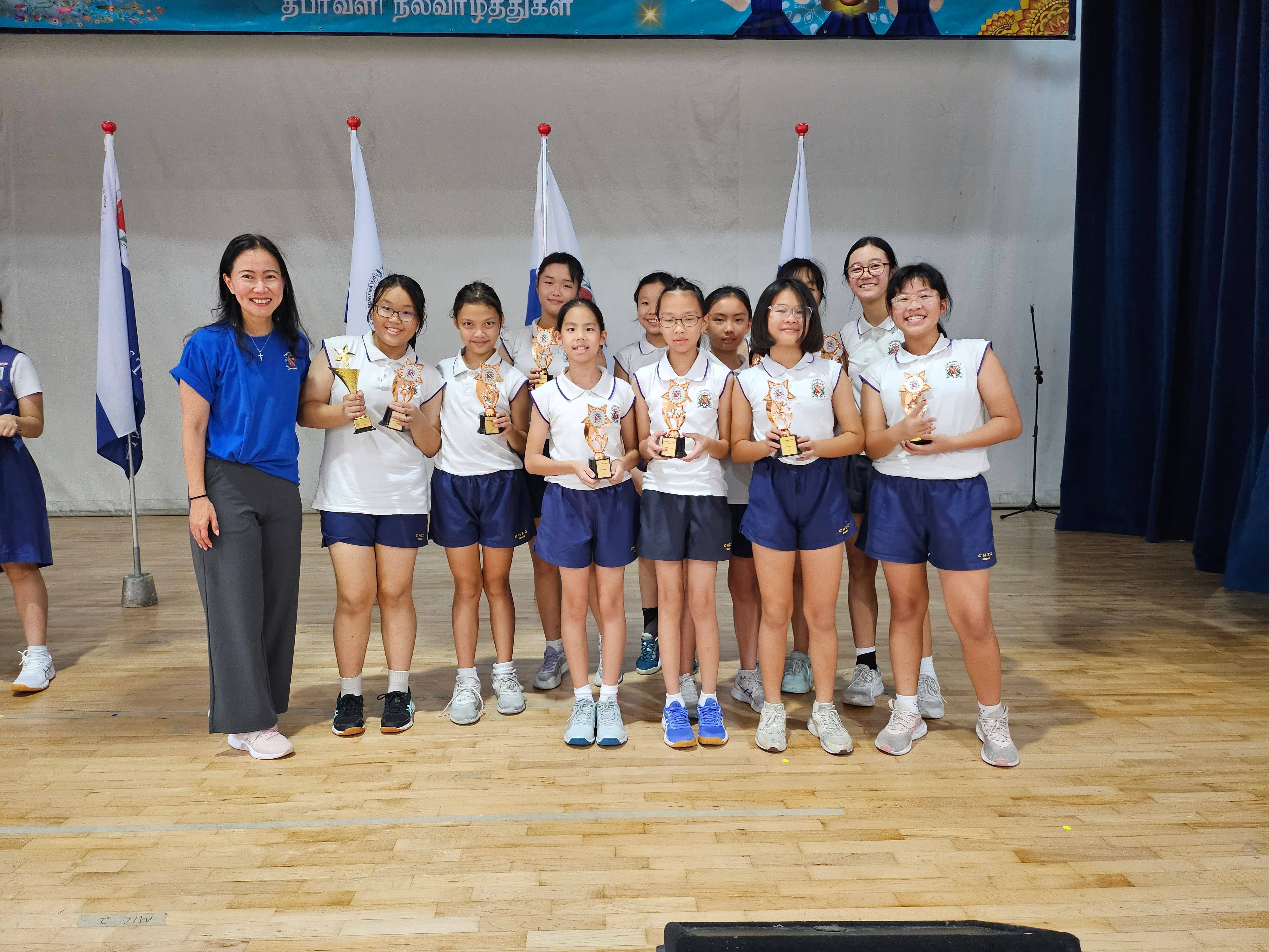 Teacher with group of students in sports uniforms on stage, each holding a trophy, flags behind them.