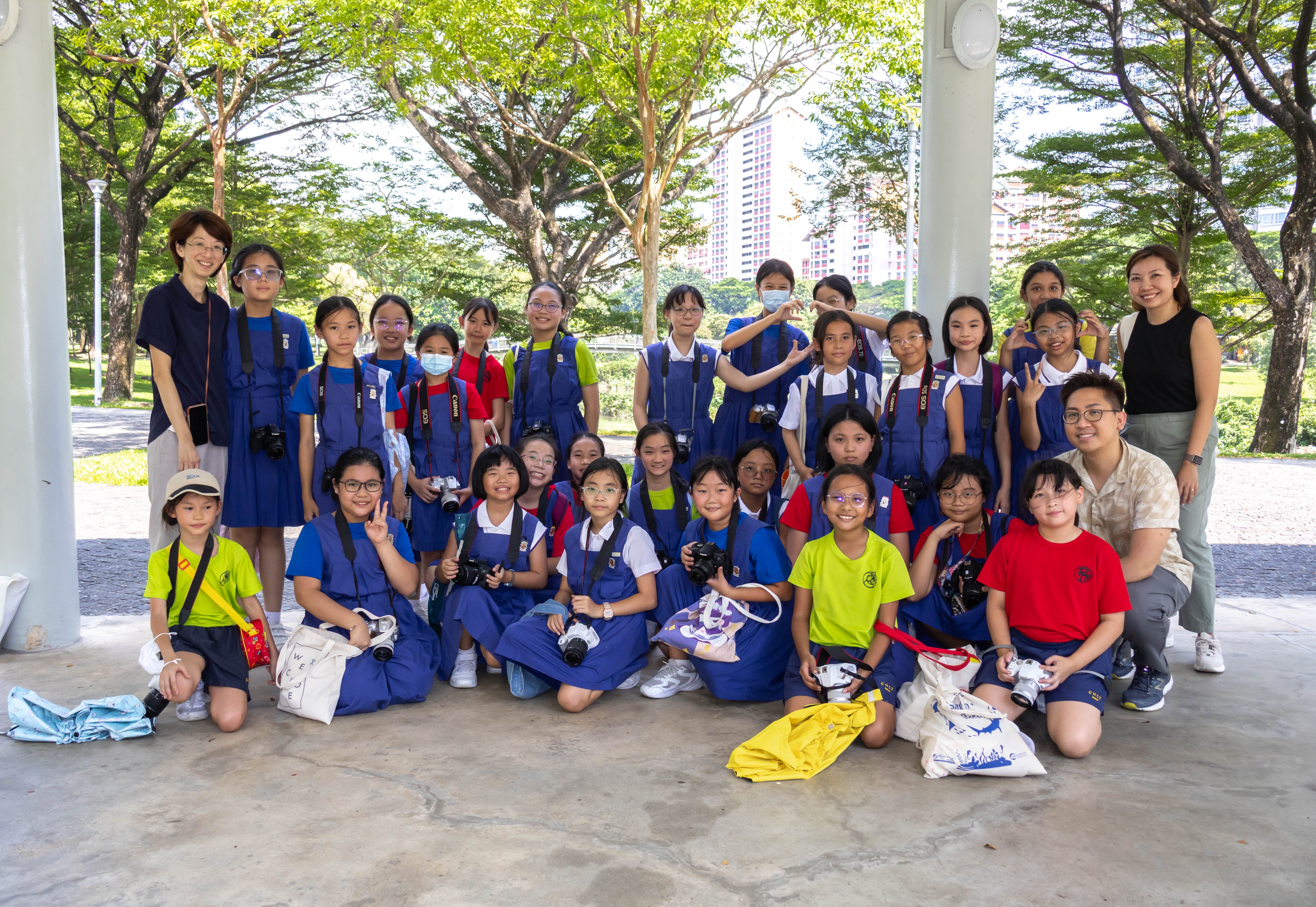 A large group of schoolchildren in blue uniforms holding cameras pose outdoors under a shaded area with trees and buildings behind.