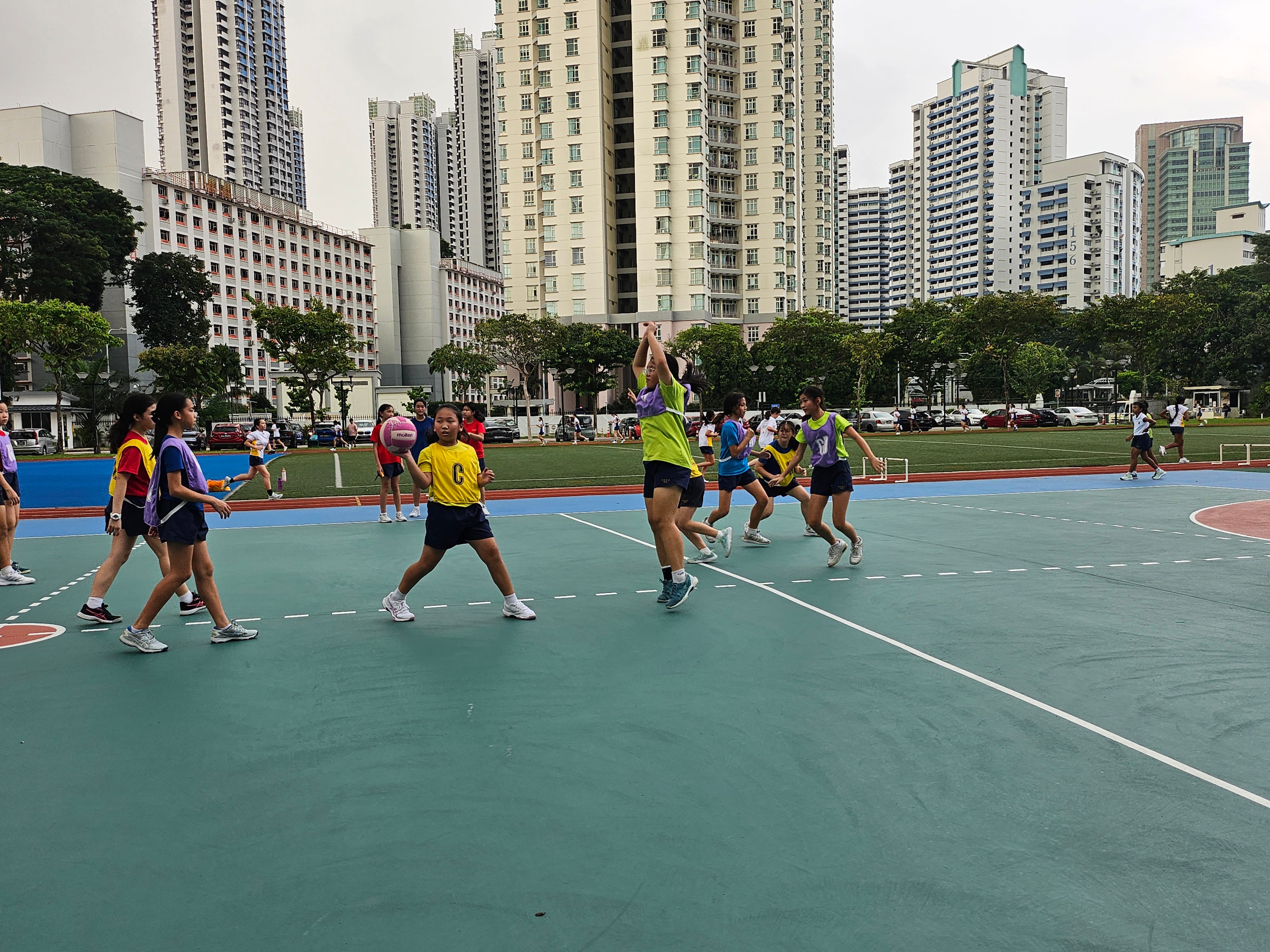 Girls in colored bibs playing netball on an outdoor court surrounded by tall city buildings and trees.