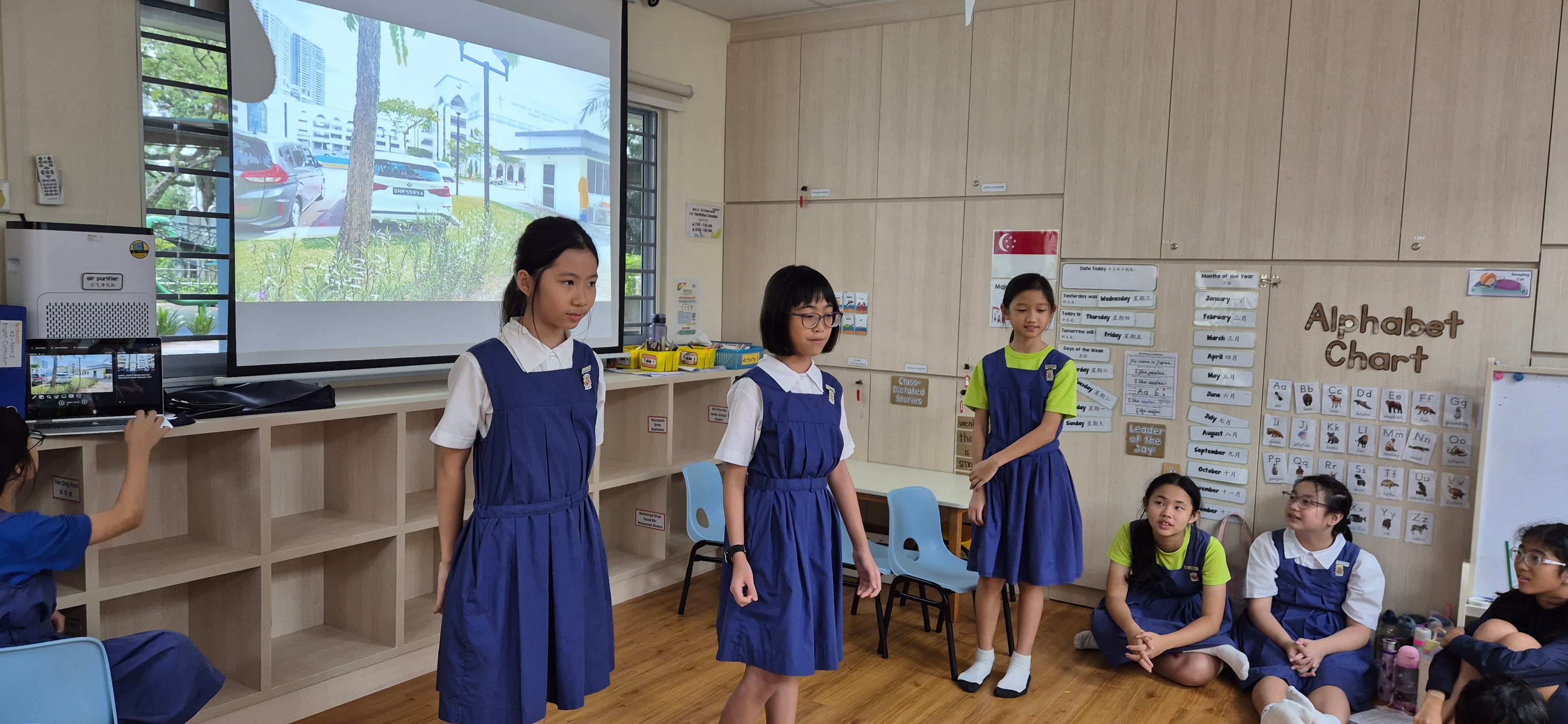 Three girls in blue uniforms stand presenting in a classroom with an alphabet chart and projected screen nearby.