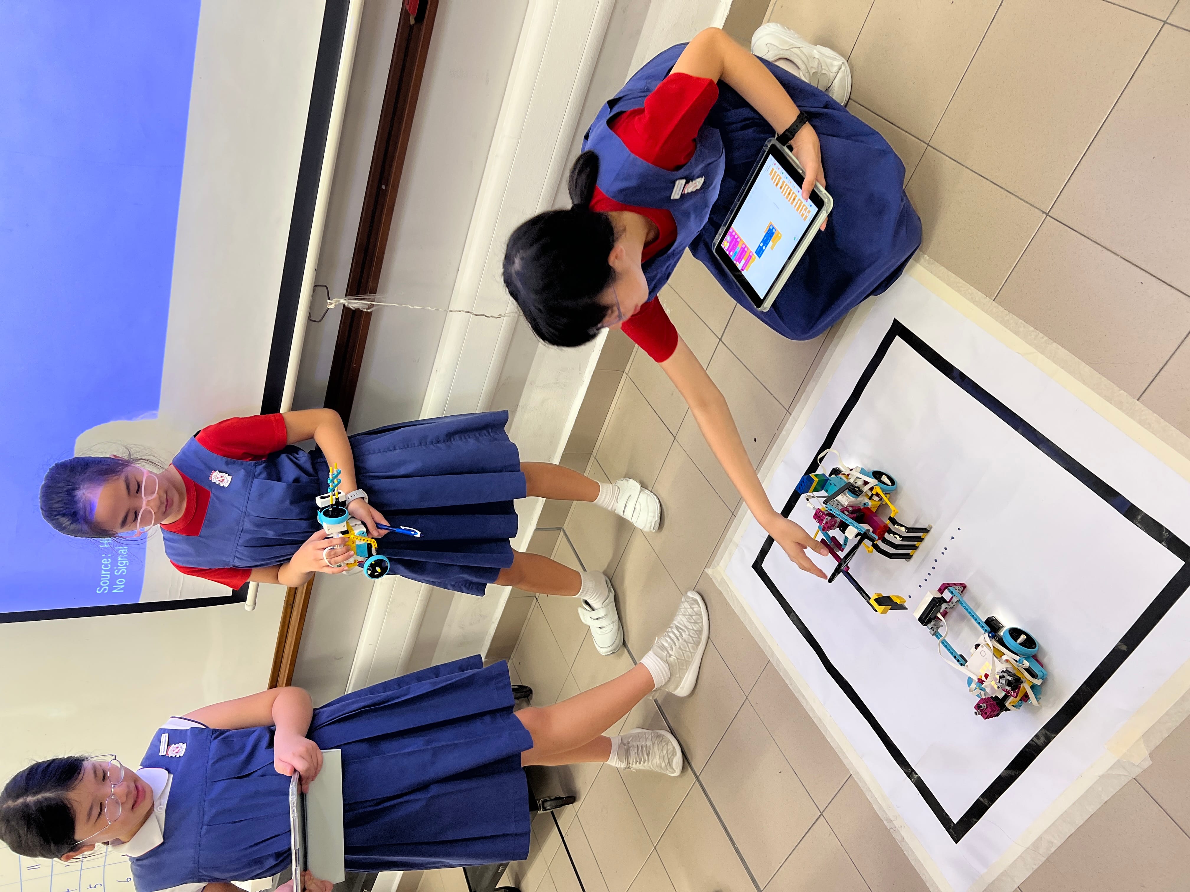 Three girls in blue school uniforms work with a LEGO robot on the floor, using tablets to program it.