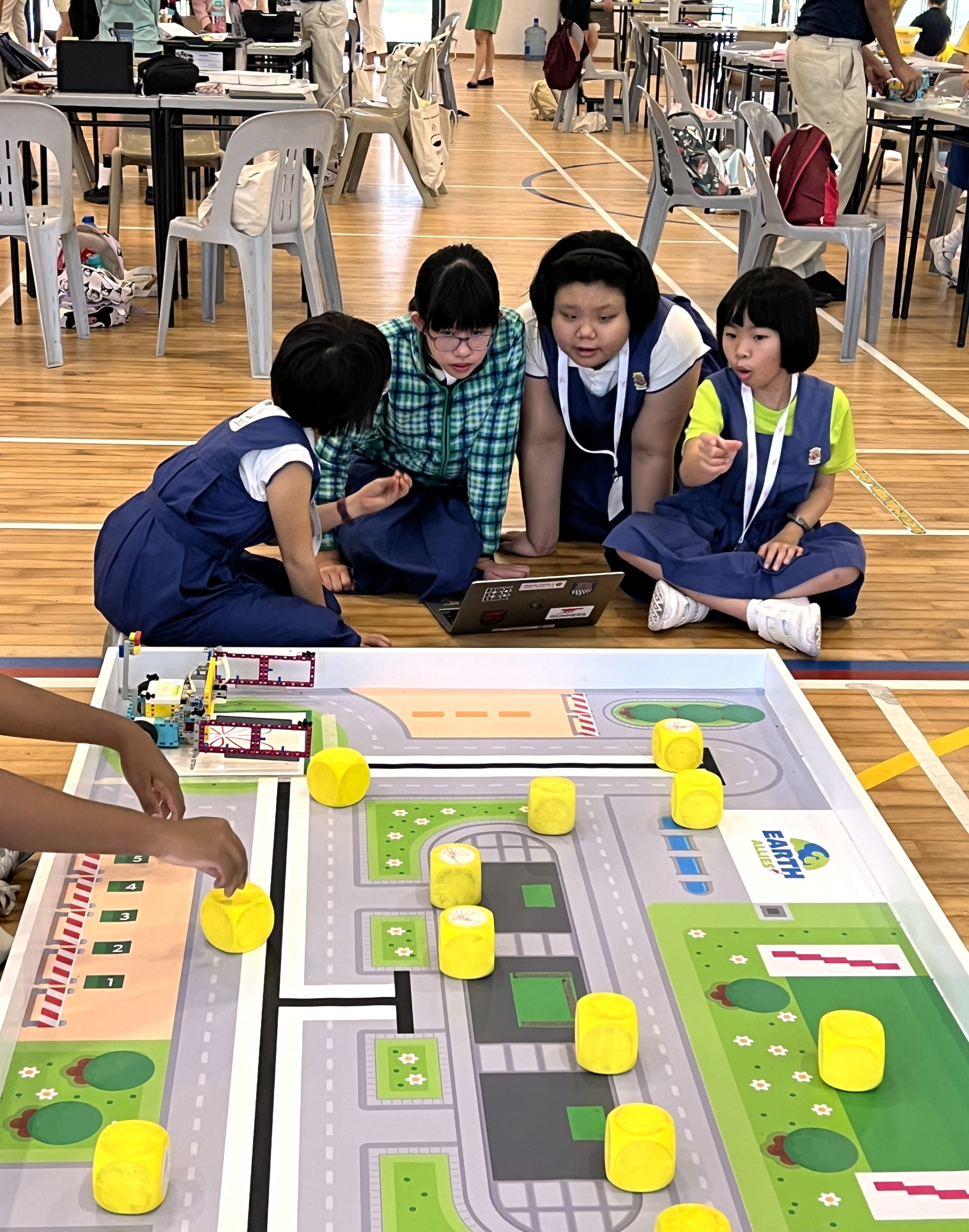 Four children in school uniforms huddle over a laptop near a city-themed robotics competition mat with yellow cylinders.