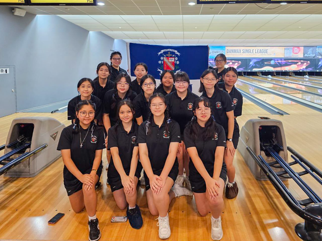 A group of young women in matching black uniforms posing together at a bowling alley with lanes visible behind them.