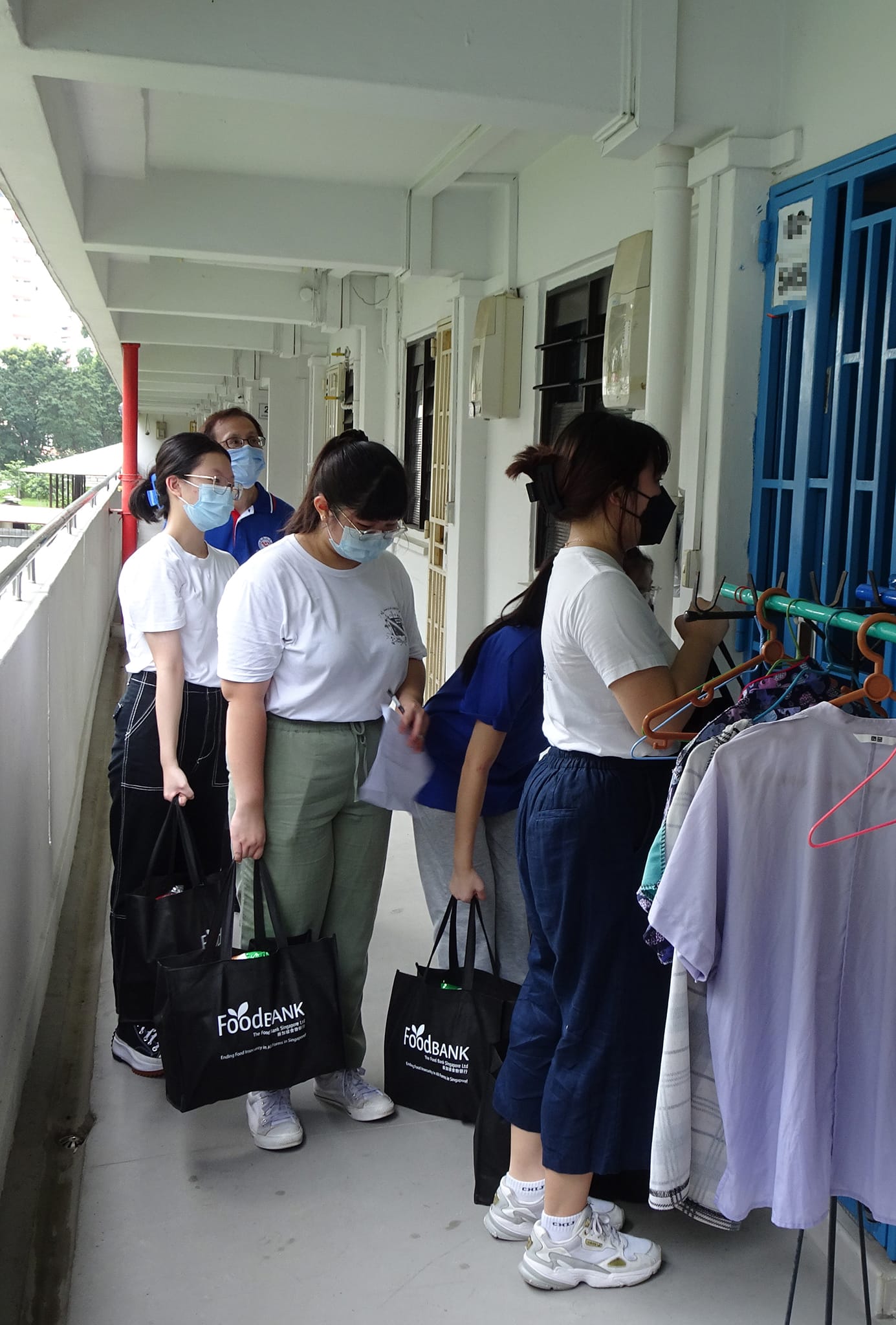 Four masked volunteers holding FoodBank bags stand in a corridor near hanging clothes outside an apartment door.