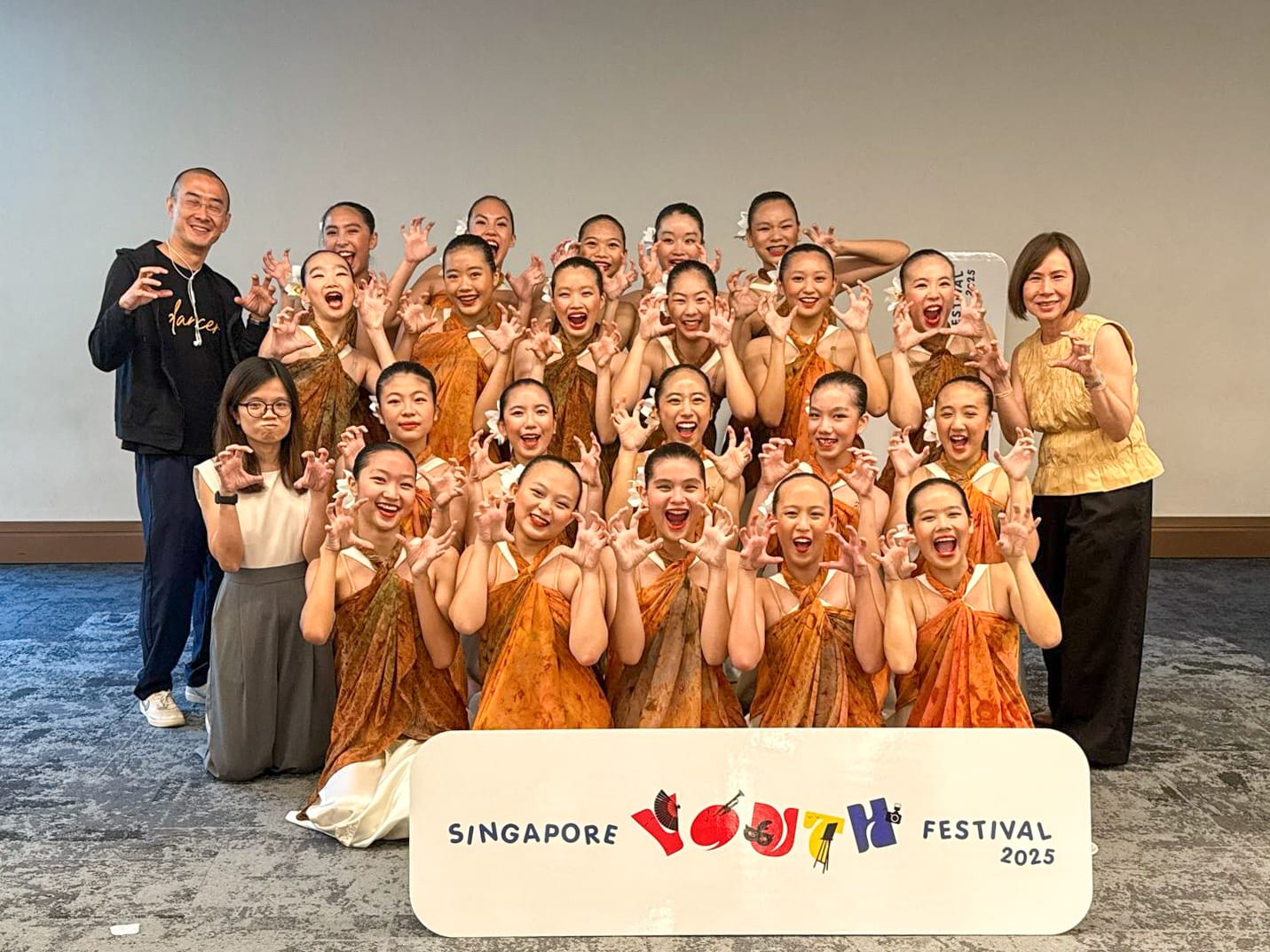Group of young dancers in orange costumes posing playfully behind a Singapore Youth Festival 2025 sign.