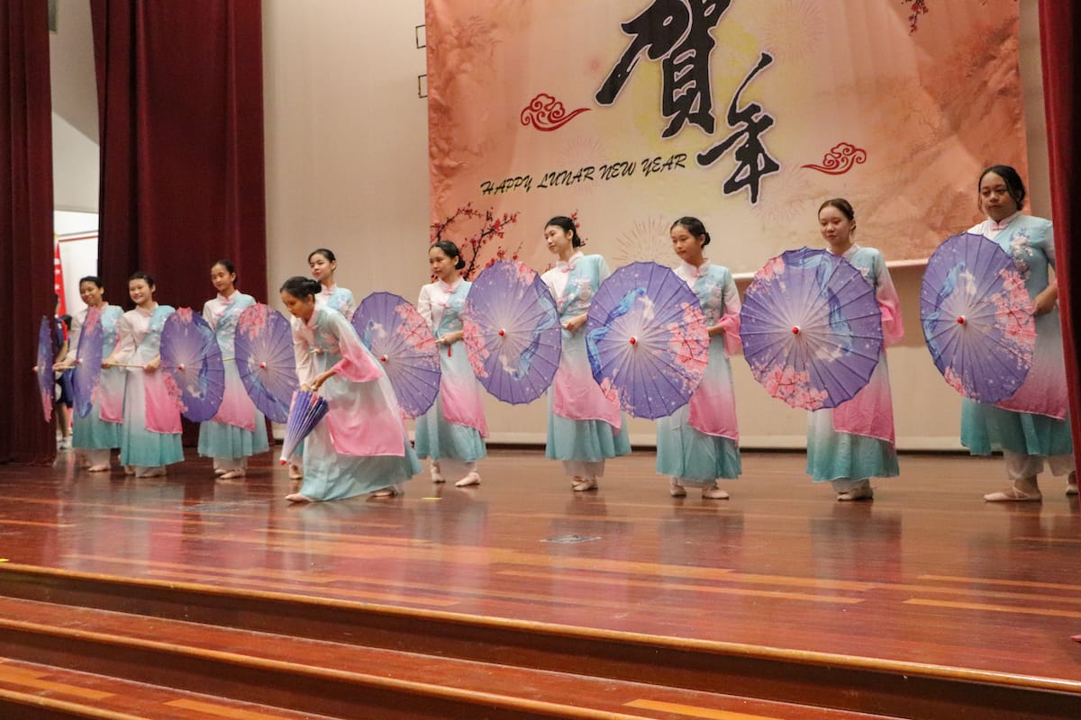 Girls in pink and teal traditional dresses hold purple parasols on stage at a Happy Lunar New Year celebration.
