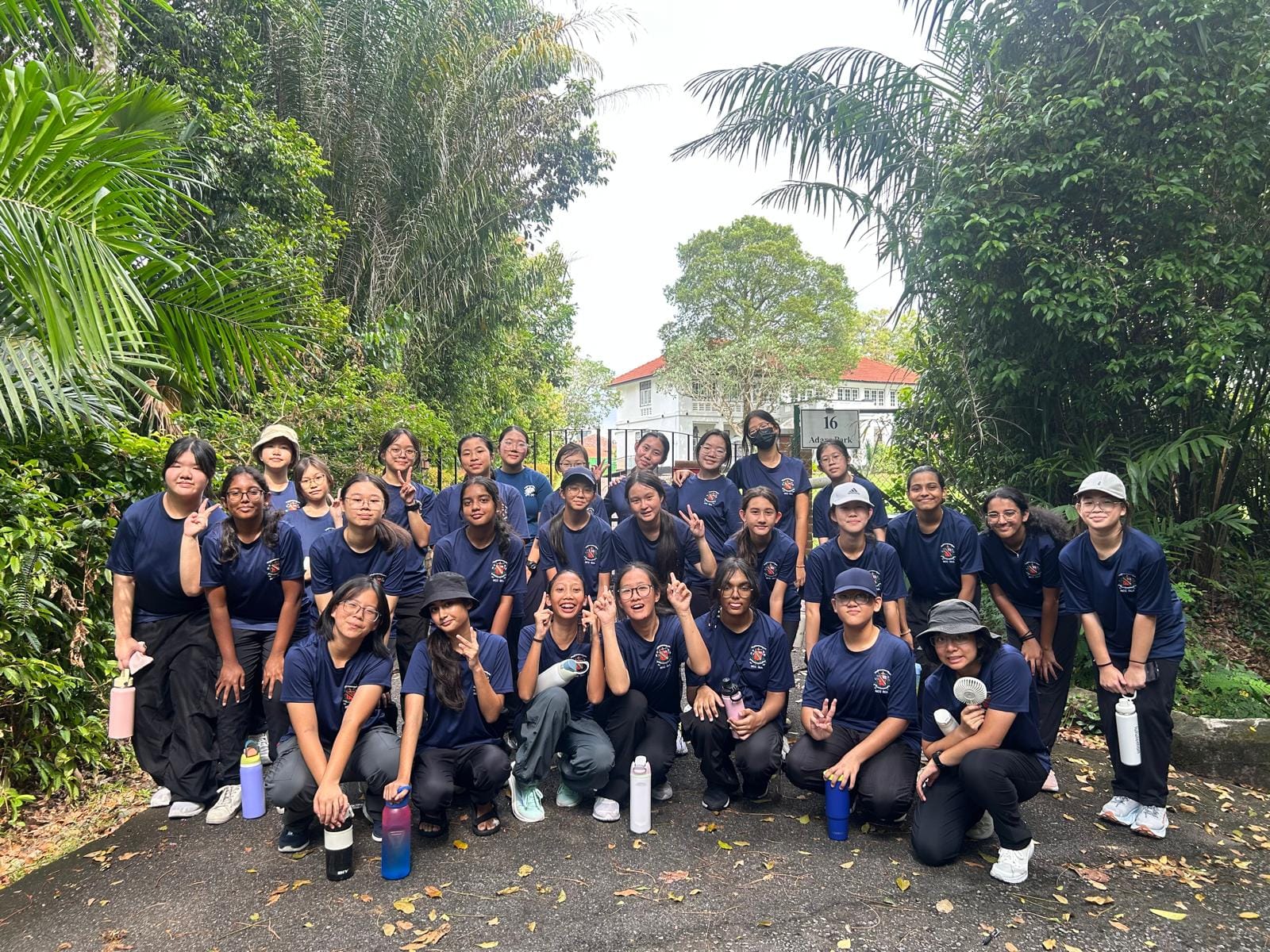 A large group of young girls in matching navy blue shirts pose outdoors near lush green trees and a gated building.