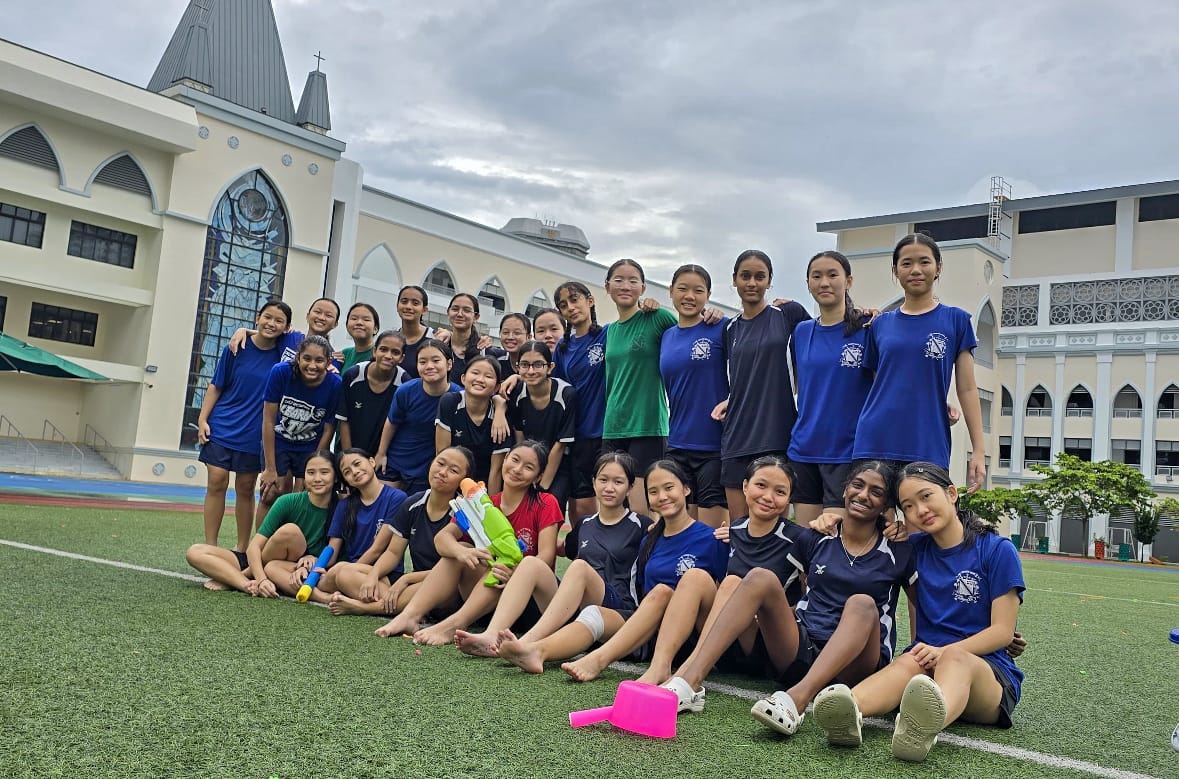 A large group of girls in blue and black sportswear pose on a green field in front of a church building.