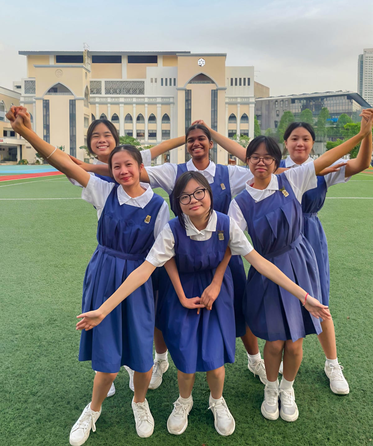 Six schoolgirls in blue pinafore uniforms pose cheerfully on a green field with a large school building behind them.