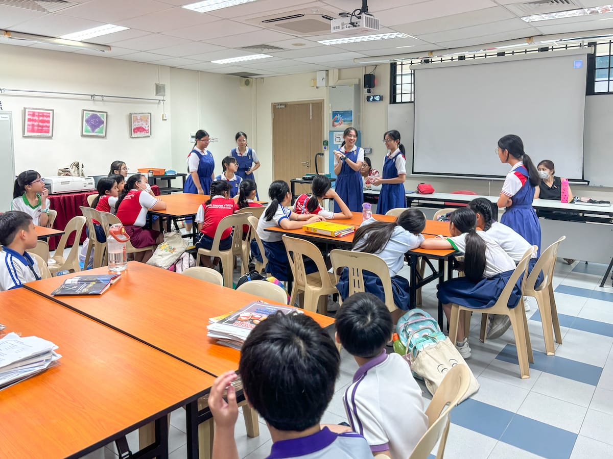 Students in uniforms sit at orange tables in a classroom while older students stand and present near a whiteboard.