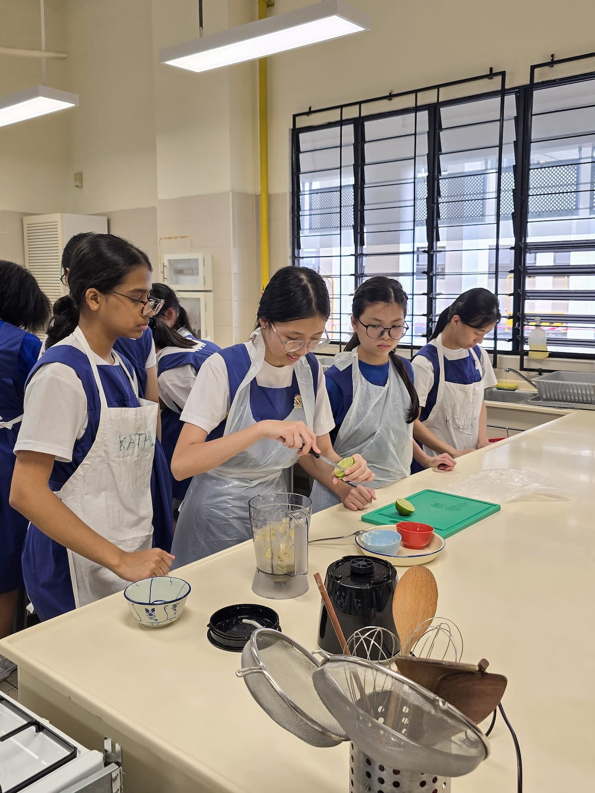 Students in blue aprons cook in a school kitchen, one cutting a lime near a blender with food inside.