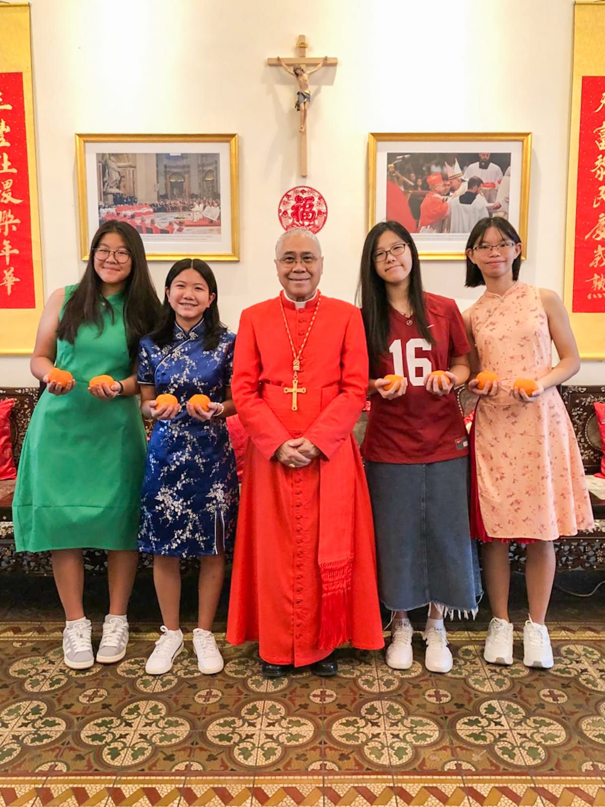 A cardinal in red robes stands with four young women holding oranges, in a room with a crucifix and Chinese decorations.