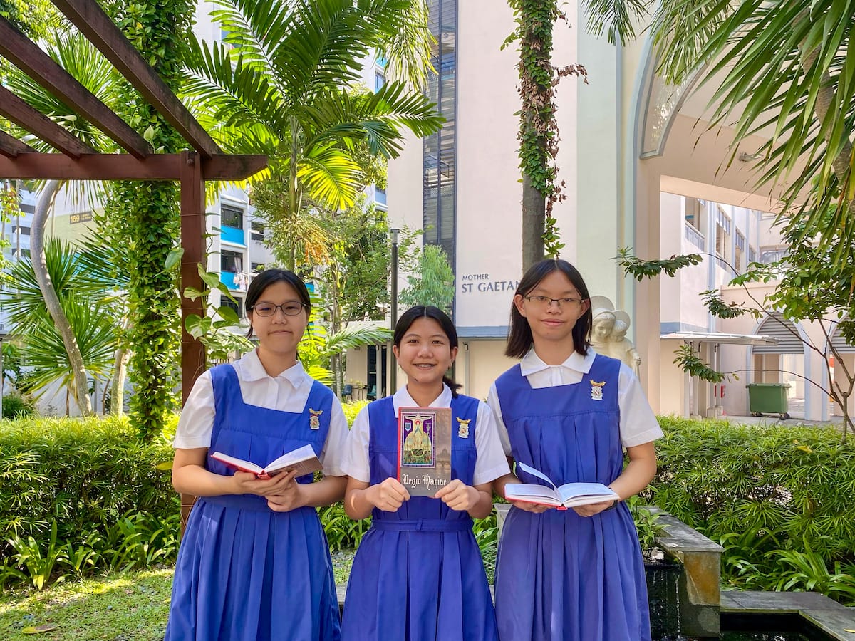 Three girls in blue school uniforms hold books outdoors near a building labeled Mother St Gaetan, surrounded by palm trees.