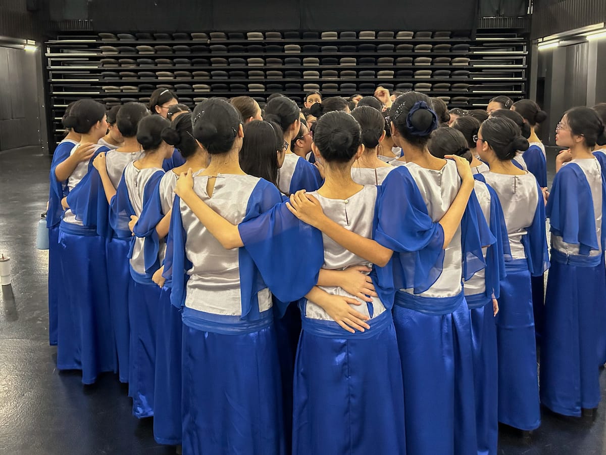 A large group of women in blue and silver gowns with hair buns stand huddled together in a circle.