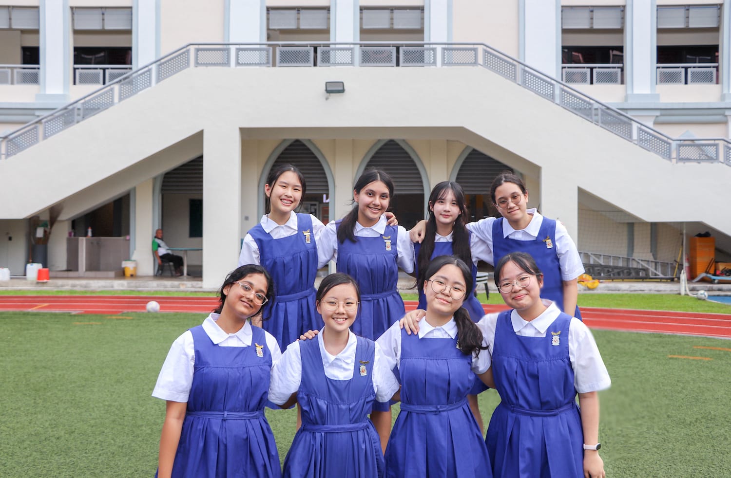 Eight schoolgirls in blue pinafore uniforms pose smiling on a green field in front of a multi-storey school building.