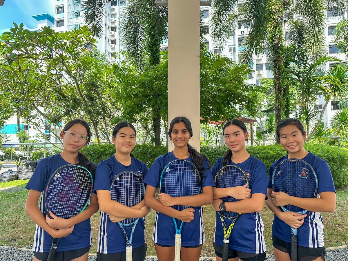 Five young girls in blue uniforms hold tennis rackets while posing outdoors near trees and buildings.