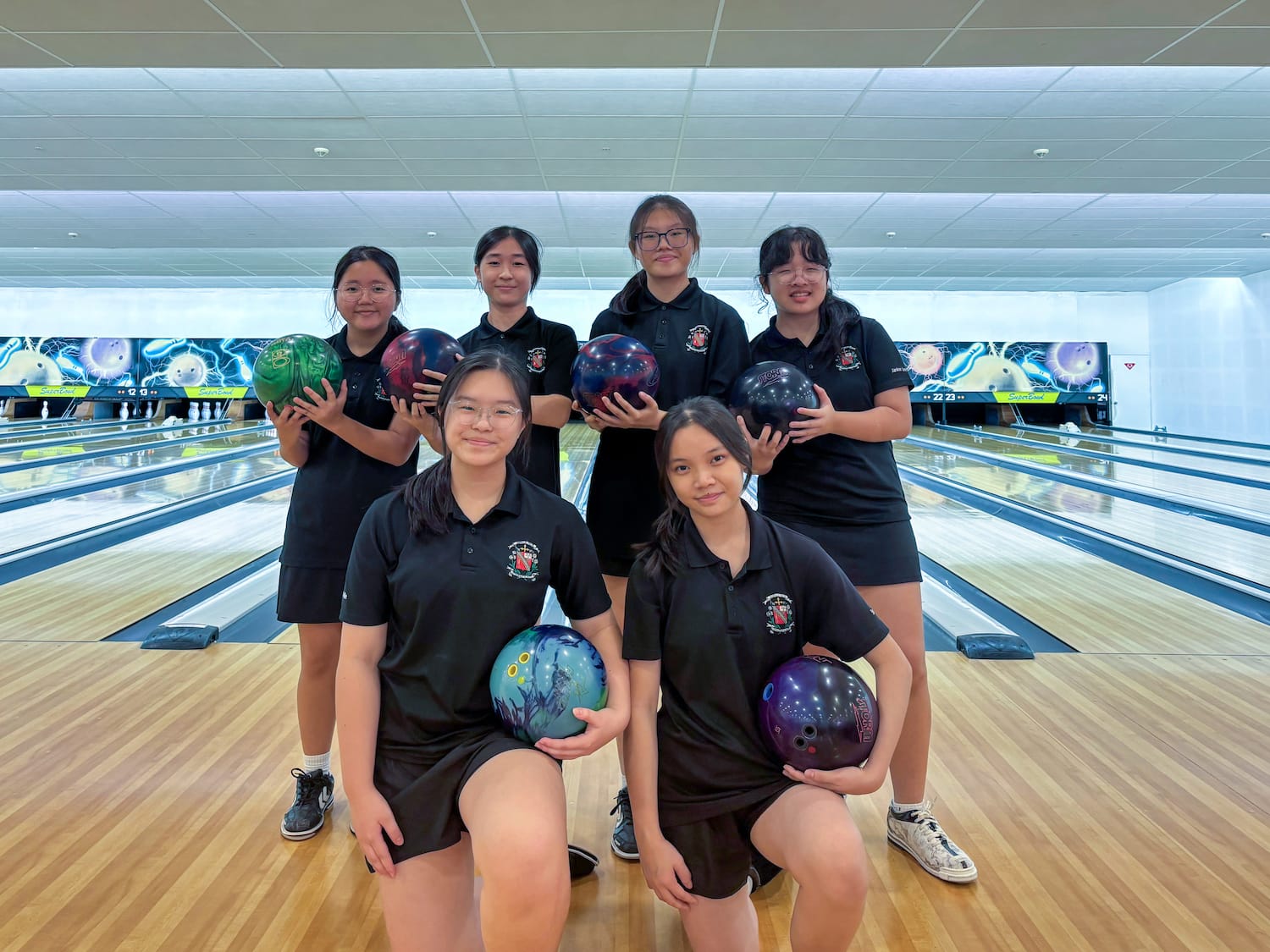 Six young women in black uniforms pose with colorful bowling balls on a bowling alley lane.