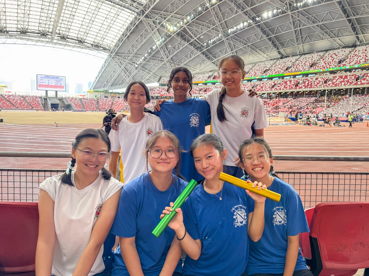 Seven girls in blue and white shirts pose in stadium stands, two holding green and yellow relay batons.