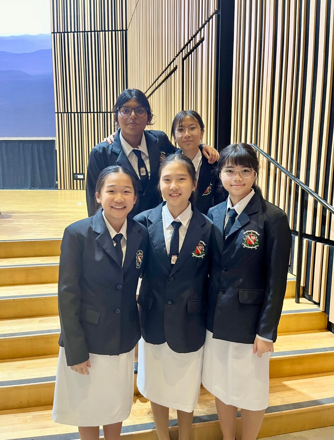 Five school girls in navy blazers, white shirts, dark ties, and white skirts pose on wooden auditorium steps.