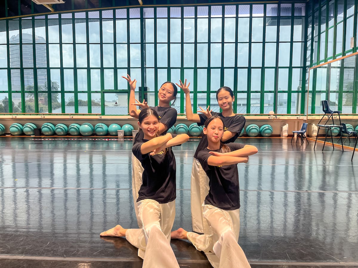 Four girls in black tops and white pants pose in dance formation on a studio floor with large green-framed windows.