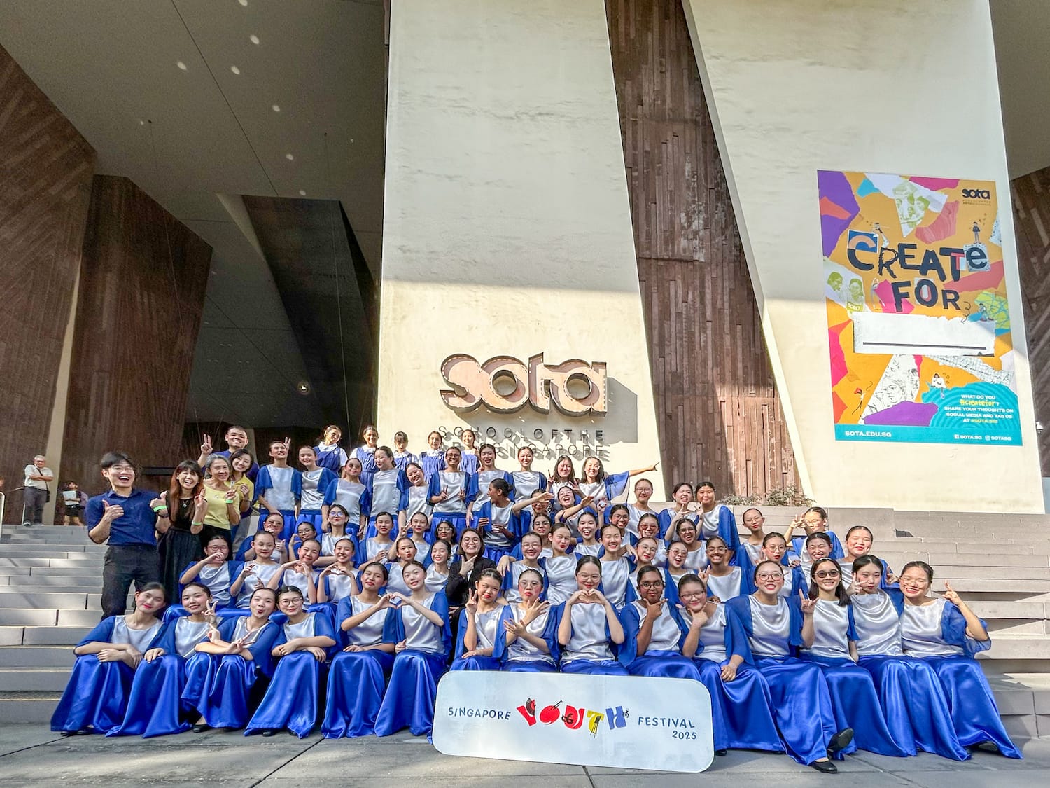 Large group of students in blue and silver choir costumes pose on SOTA steps with a Singapore Youth Festival 2025 sign.