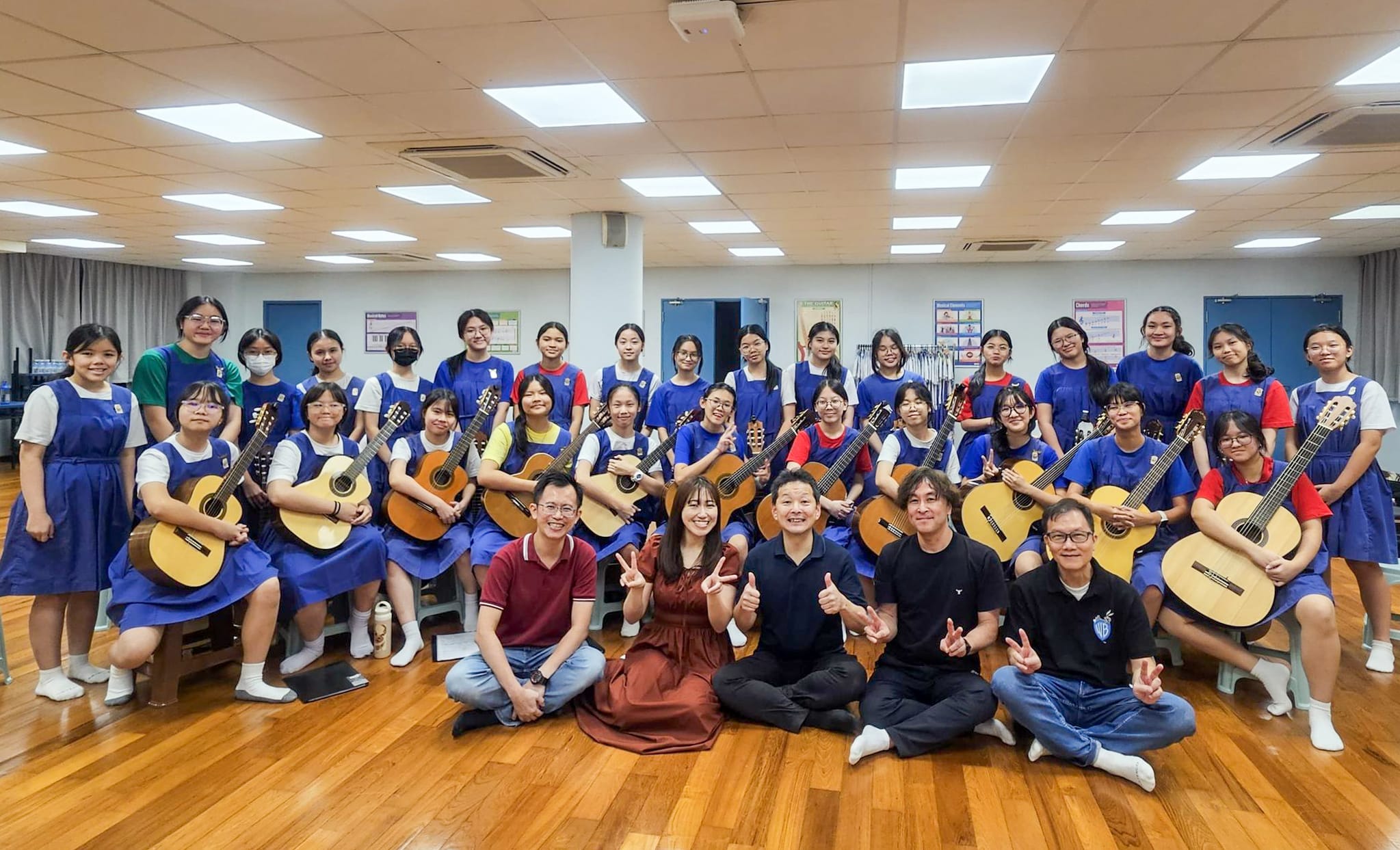 A large group of students in blue uniforms holding acoustic guitars pose with five adults in a school music room.