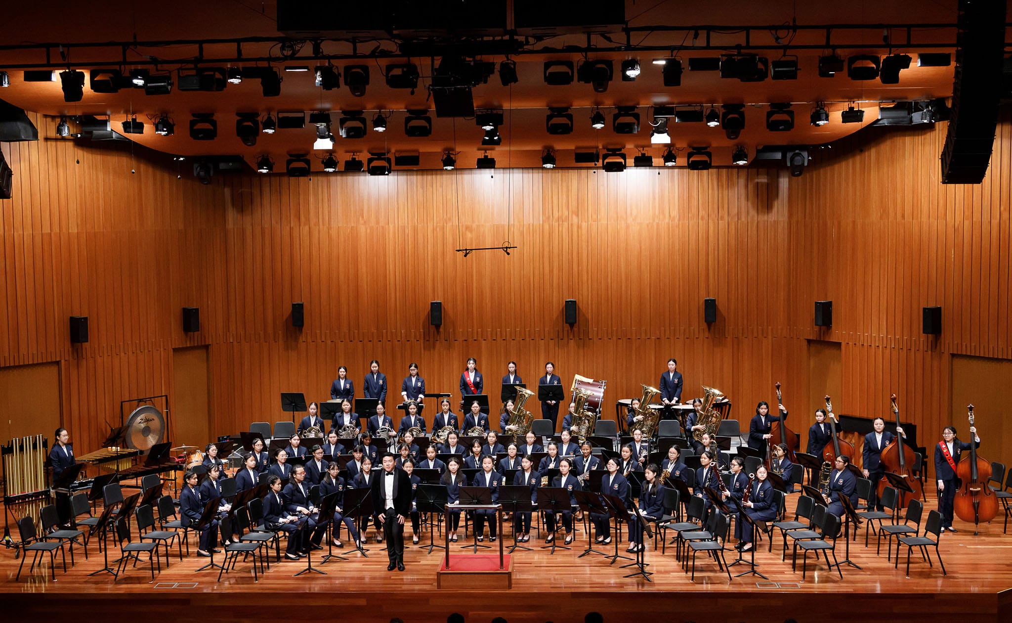 Large youth wind band in navy uniforms on a wooden concert hall stage, with a conductor standing at center front.