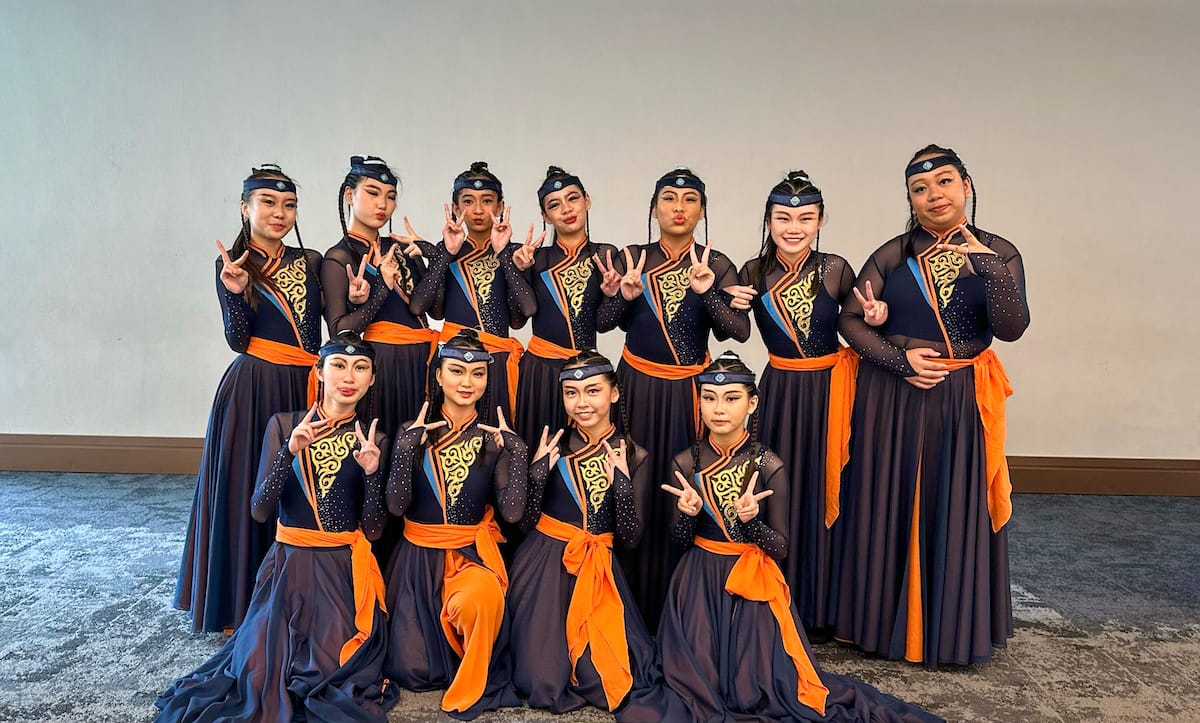 Eleven female dancers in matching navy and orange traditional costumes with gold embroidery pose with peace signs.
