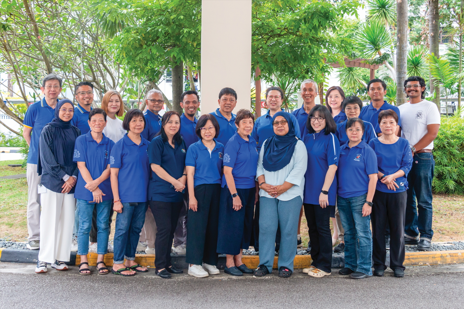 A large group of about 20 adults, most wearing blue polo shirts, posing together outdoors near trees.
