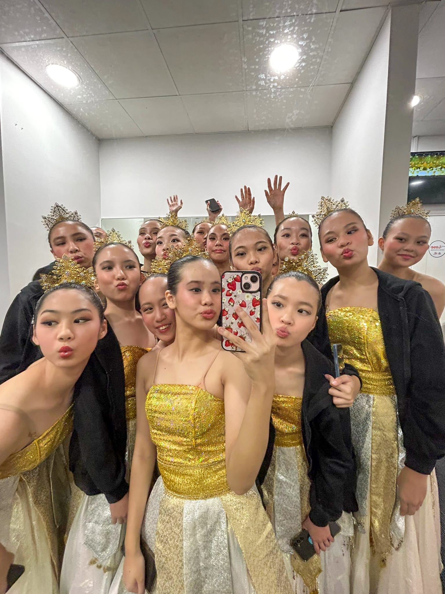 Group of young female dancers in gold costumes and ornate crowns taking a mirror selfie, posing with duck faces.