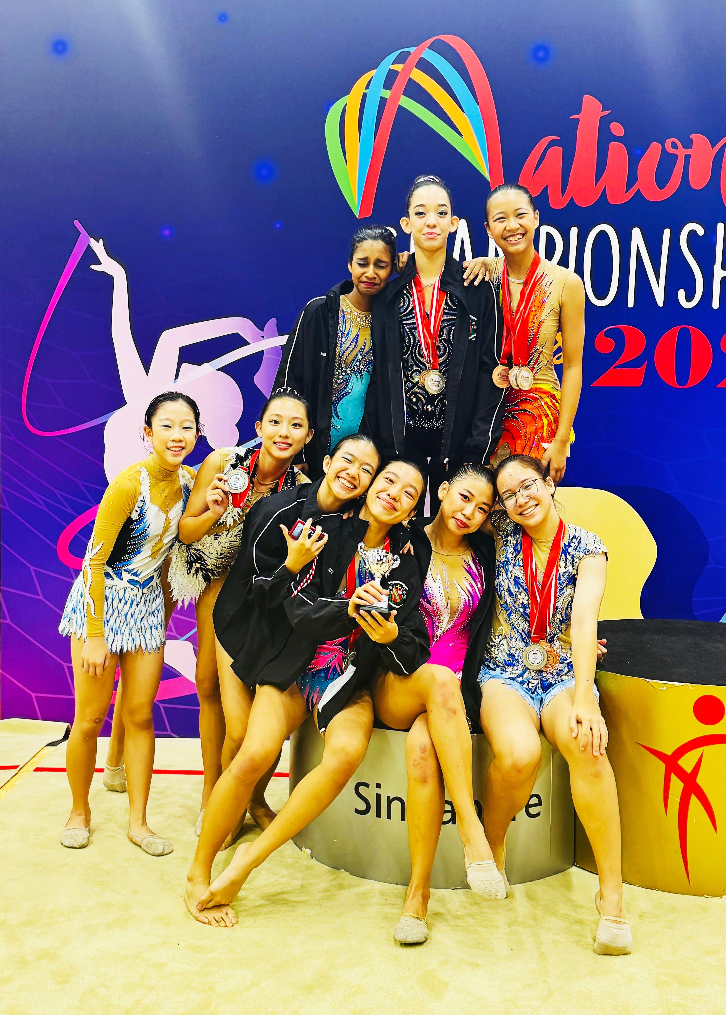 Eight young female gymnasts pose together on a podium at the Singapore National Championship, some wearing medals.