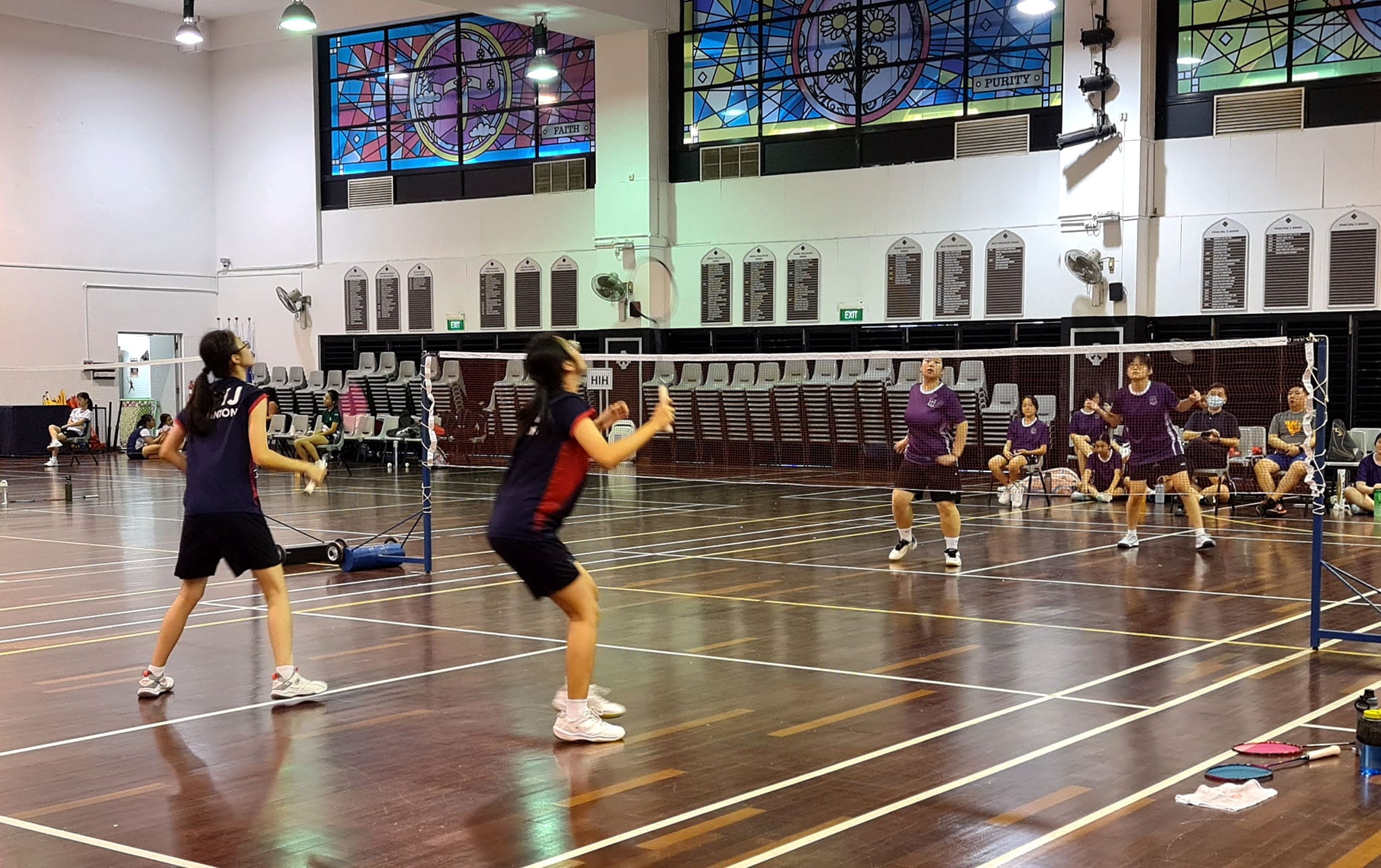 Four players in two teams play badminton indoors in a hall with colorful stained glass windows above.