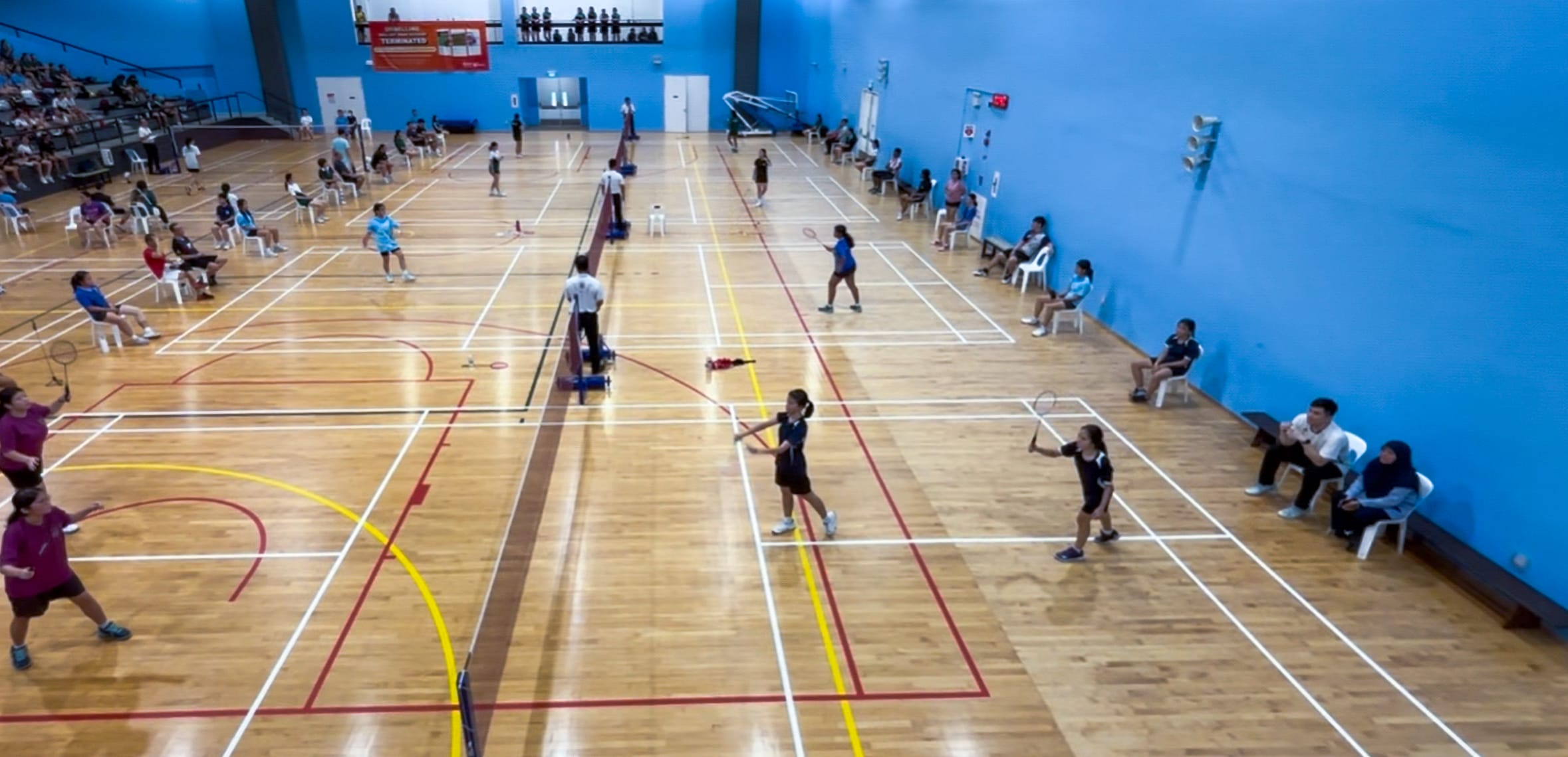 Multiple badminton matches being played simultaneously on a wood-floored indoor court with blue walls and spectators seated along the sides.