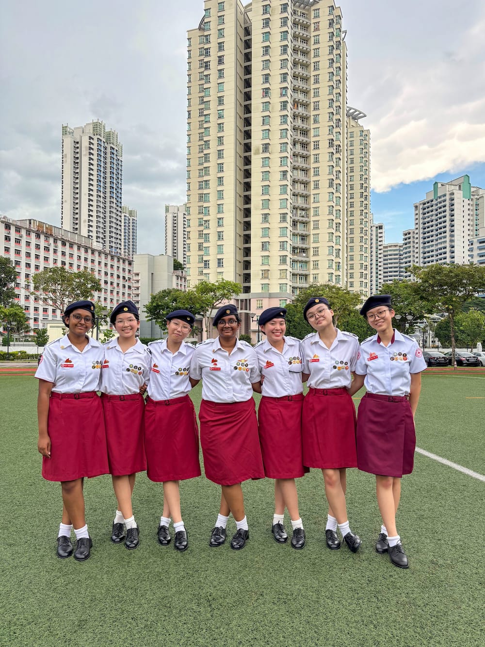 Seven girls in white shirts, red skirts, and navy berets stand smiling on a green field with tall buildings behind them.