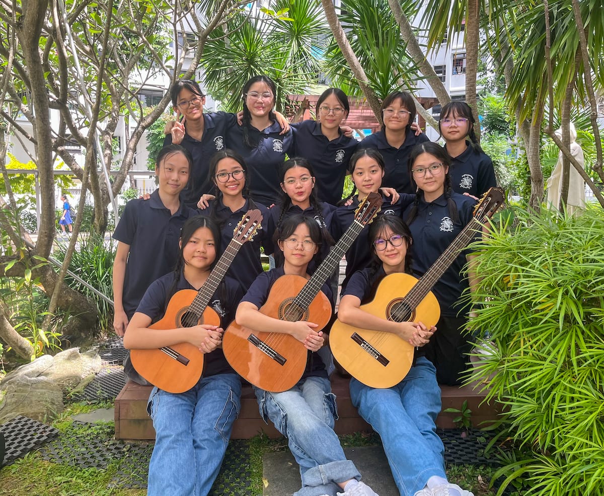 A group of young girls in navy uniforms pose outdoors, with three in front holding acoustic guitars.
