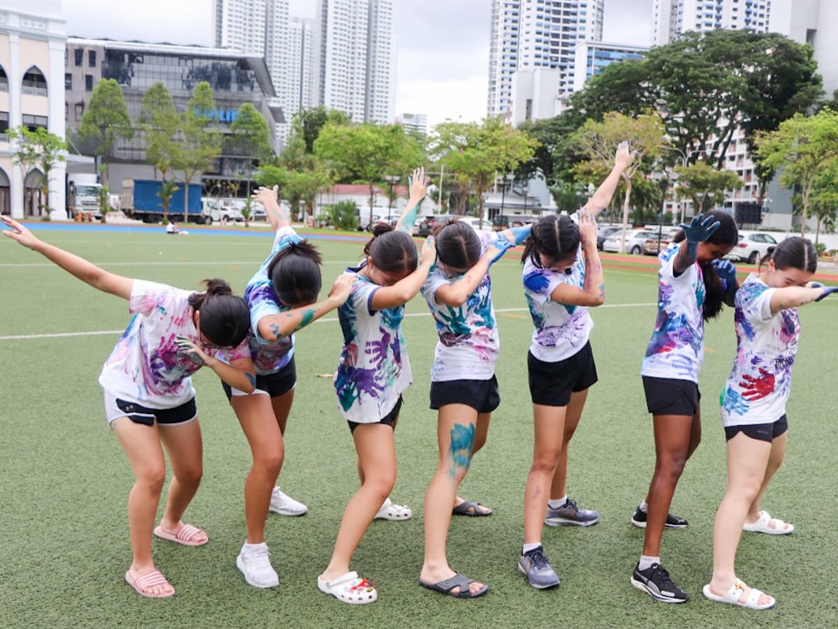 Seven girls in paint-covered white shirts strike a dabbing pose on a green field with city buildings behind them.