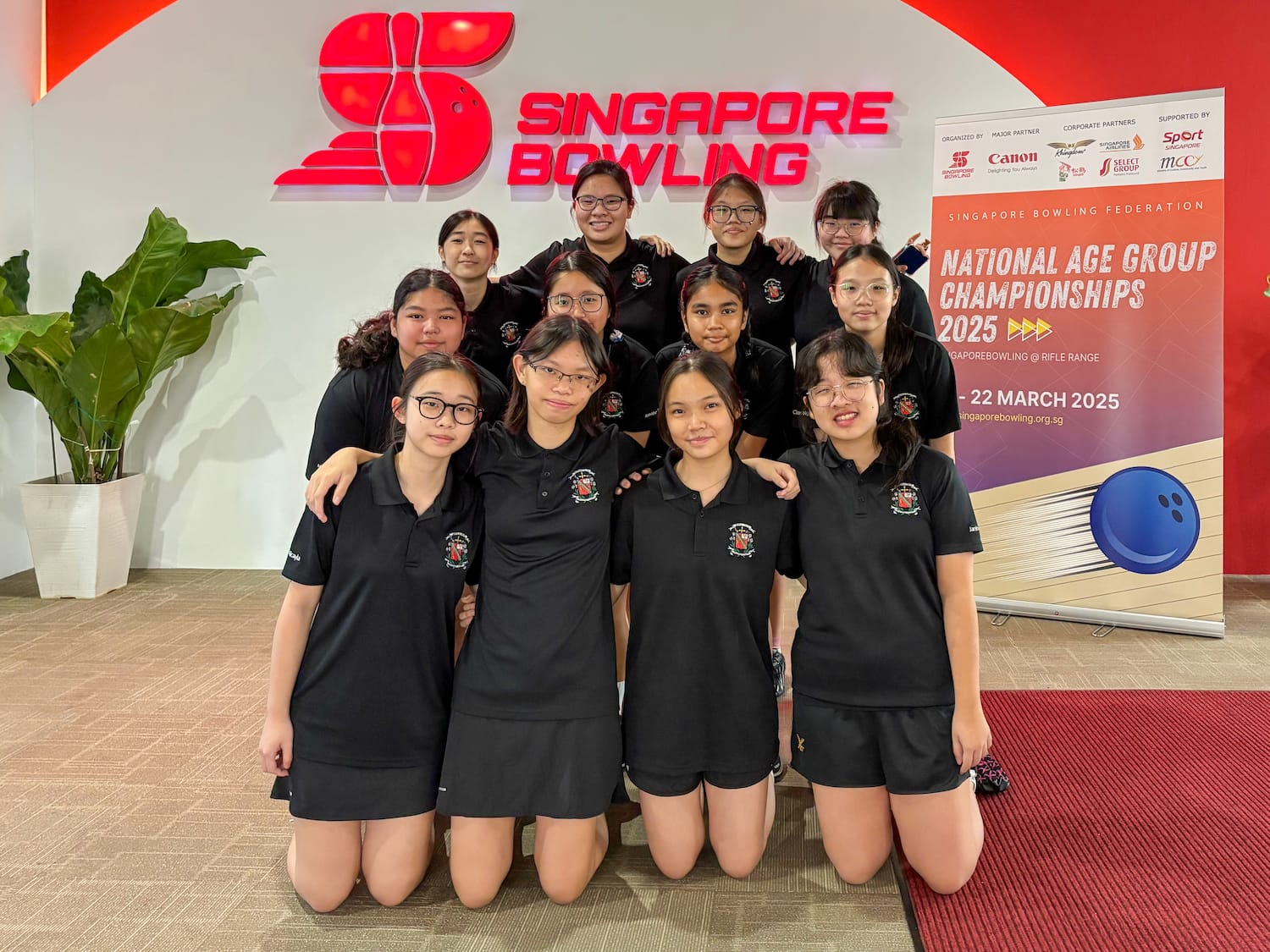 A group of young girls in black uniforms pose at Singapore Bowling's National Age Group Championships 2025 banner.