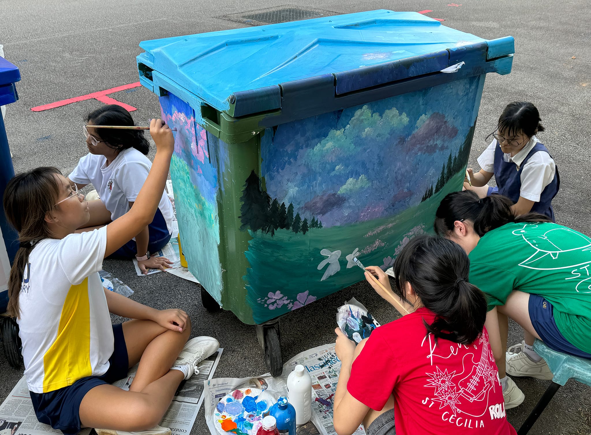 Several students sit outdoors painting a colorful nature mural of trees and sky on a large green bin.