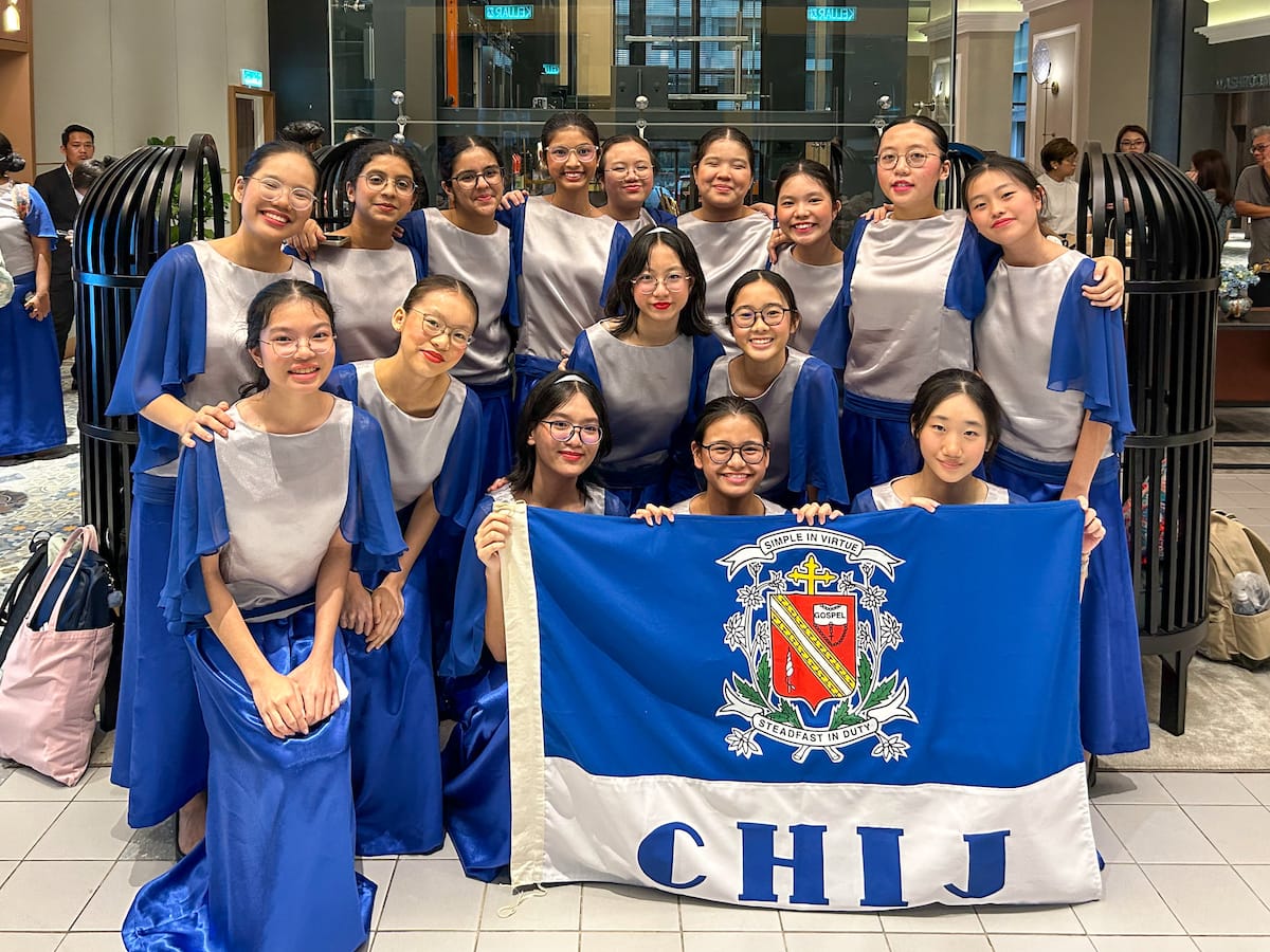 A group of girls in blue and white choir uniforms pose together holding a blue CHIJ school flag.