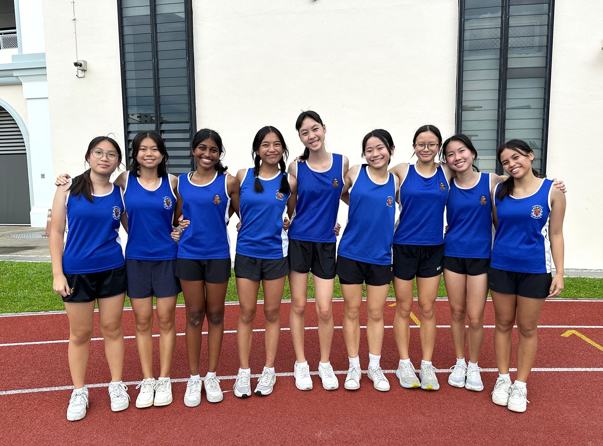Eight young women in matching blue athletic jerseys stand together smiling on a red running track.