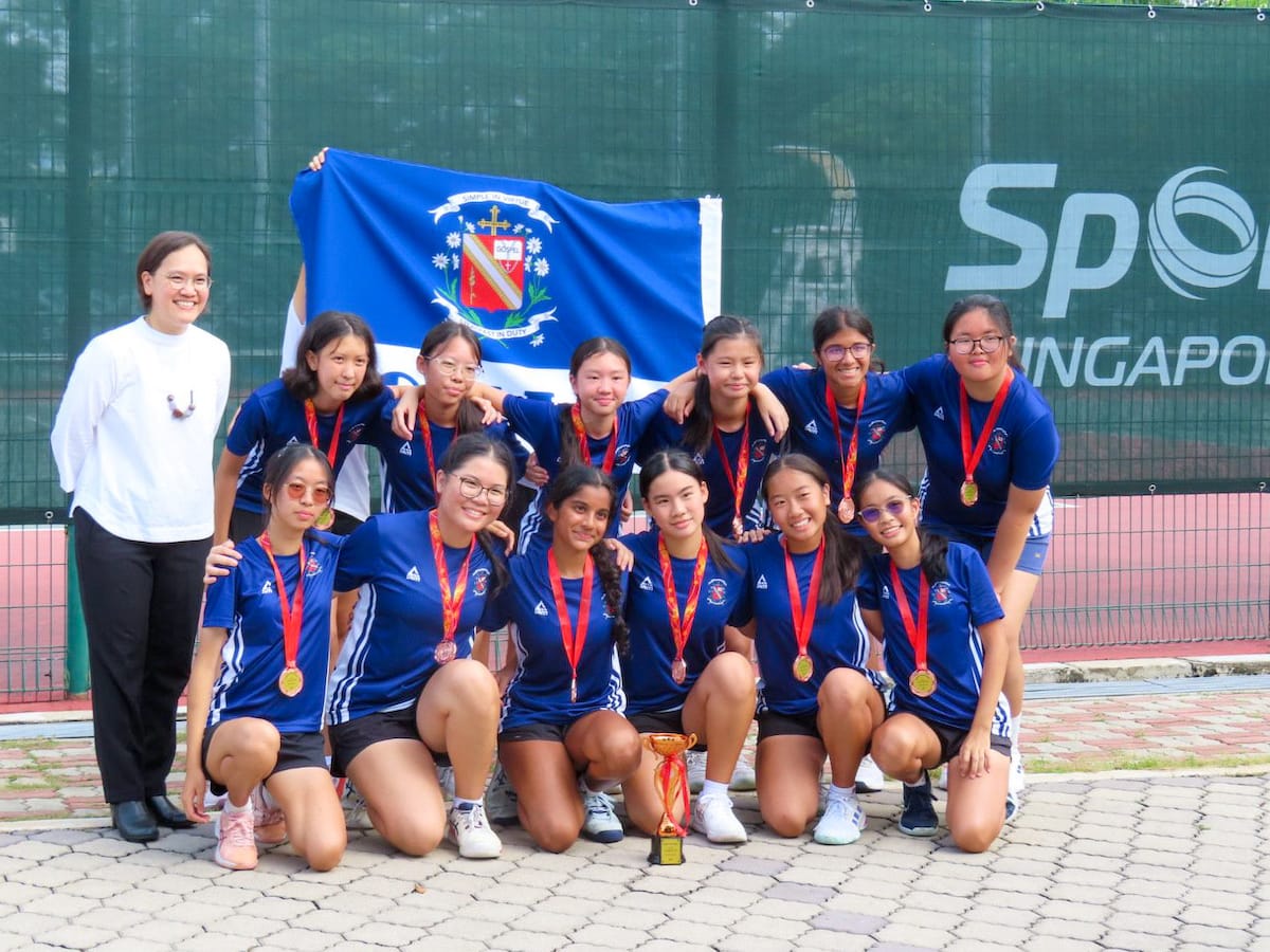 A girls sports team in blue jerseys poses with gold medals, a trophy, and a blue school flag outdoors.