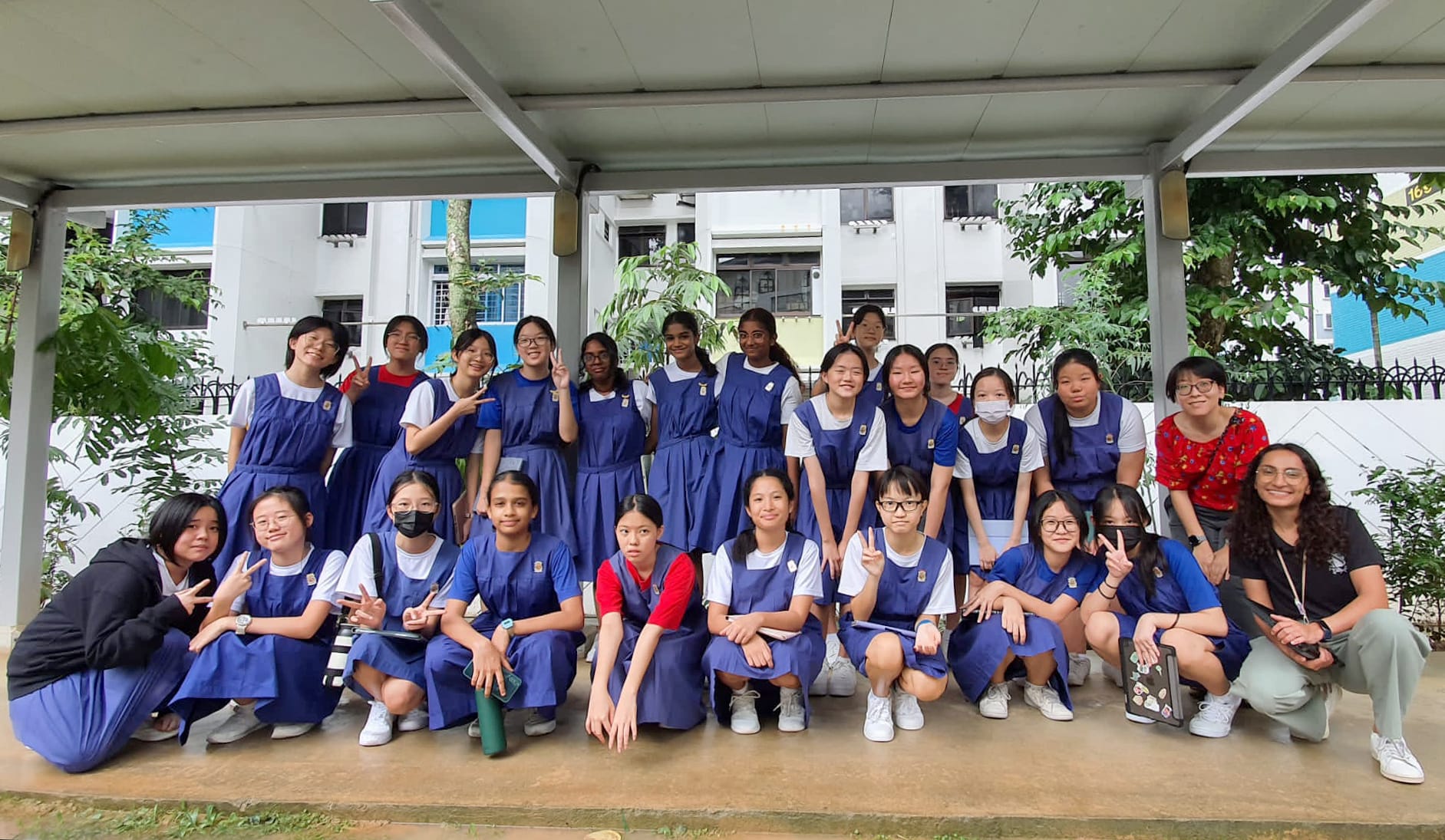 A large group of students in blue uniforms pose outdoors under a covered area, many making peace signs.