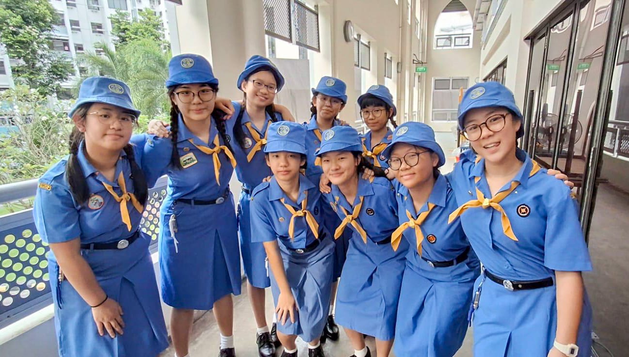 Nine young girls in blue scout uniforms with yellow neckerchiefs and blue caps pose together in a school corridor.