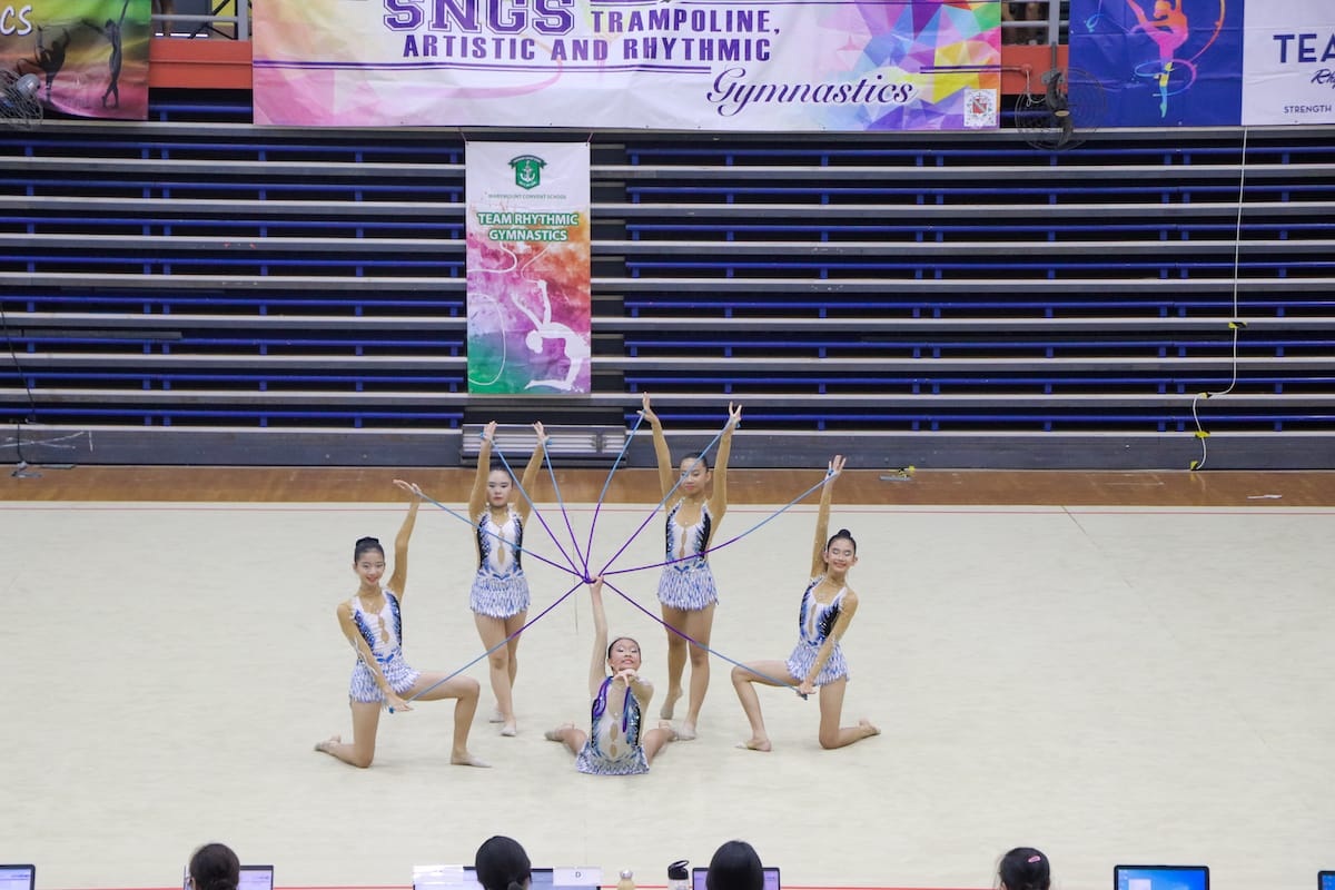 Five rhythmic gymnasts in matching leotards perform a group routine with ropes forming a star pattern on a competition floor.