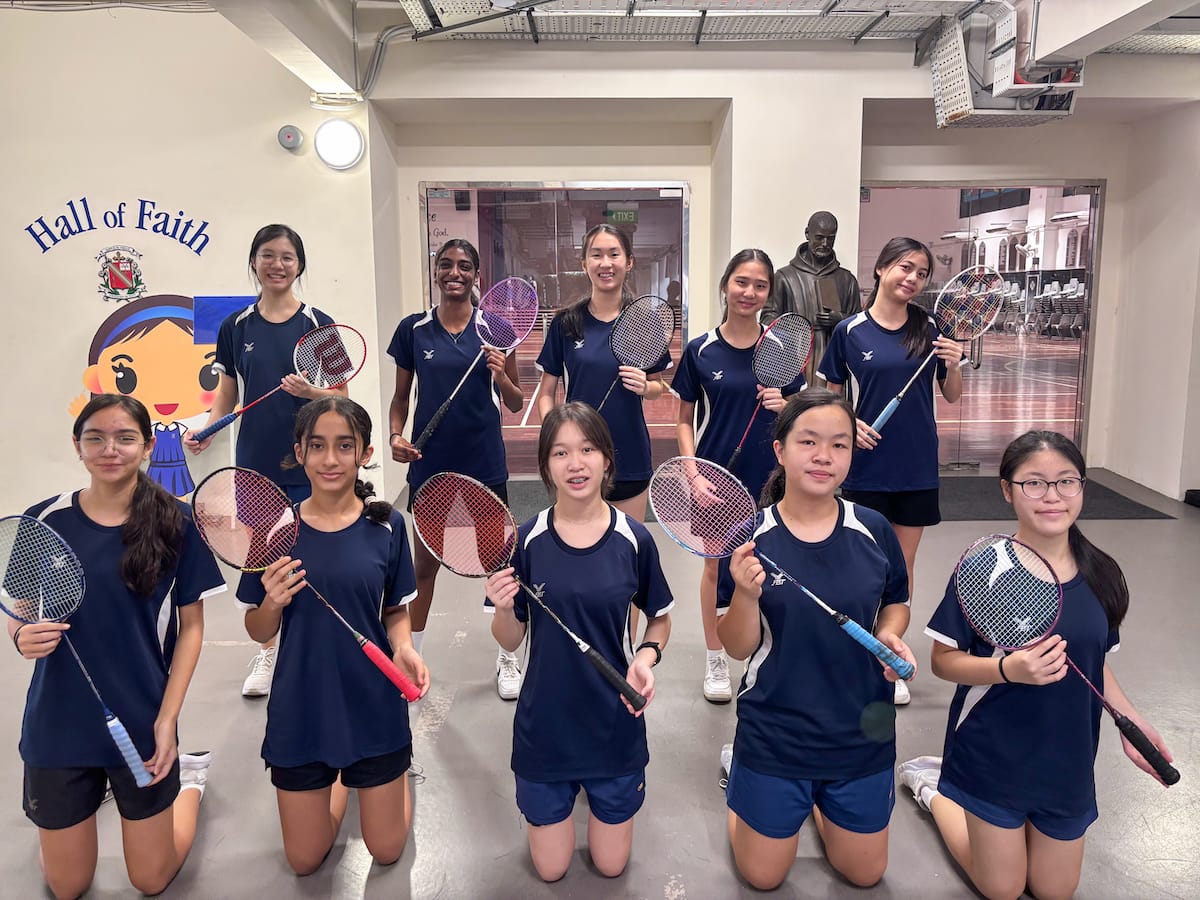 Eleven girls in navy uniforms pose in two rows holding badminton rackets indoors near a Hall of Faith sign.