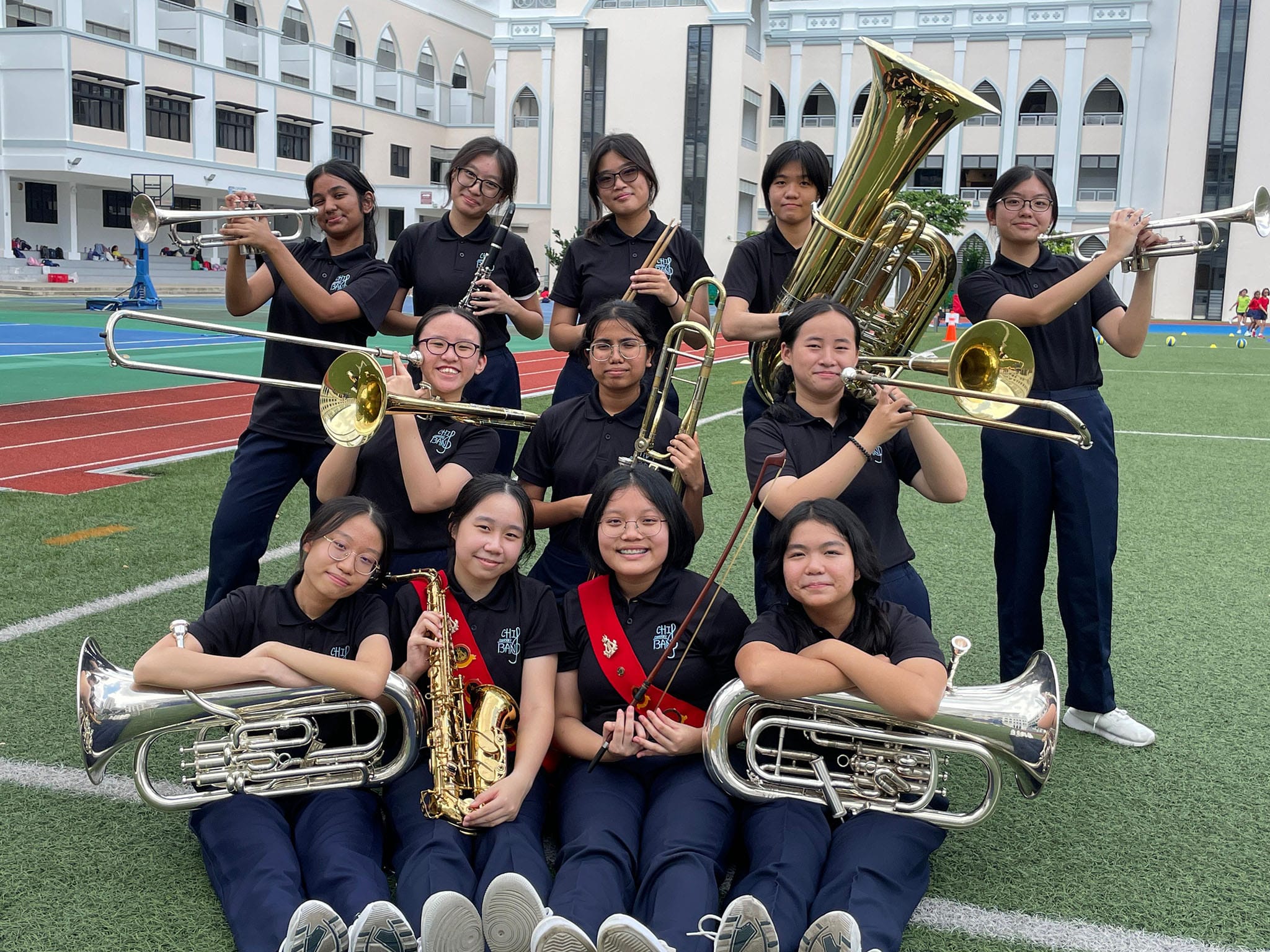 A group of school band students in navy uniforms pose with brass instruments on a sports field.