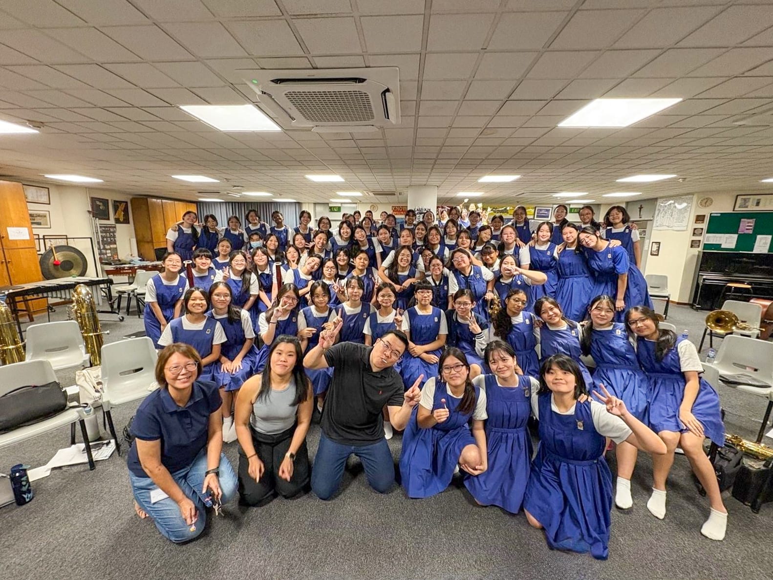 Large group of students in blue uniforms posing with two adults in a music room with instruments visible.