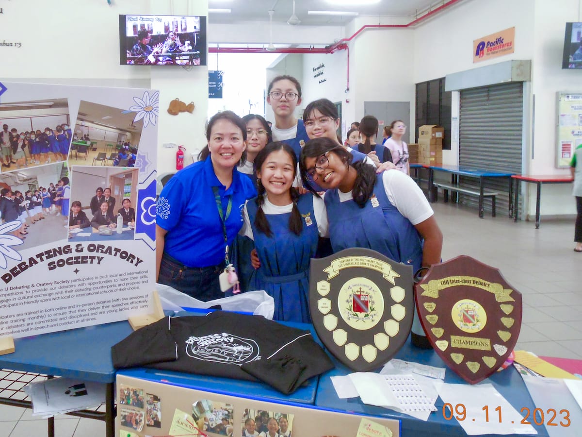 A teacher and students pose at a Debating and Oratory Society booth with two shield trophies on the table.