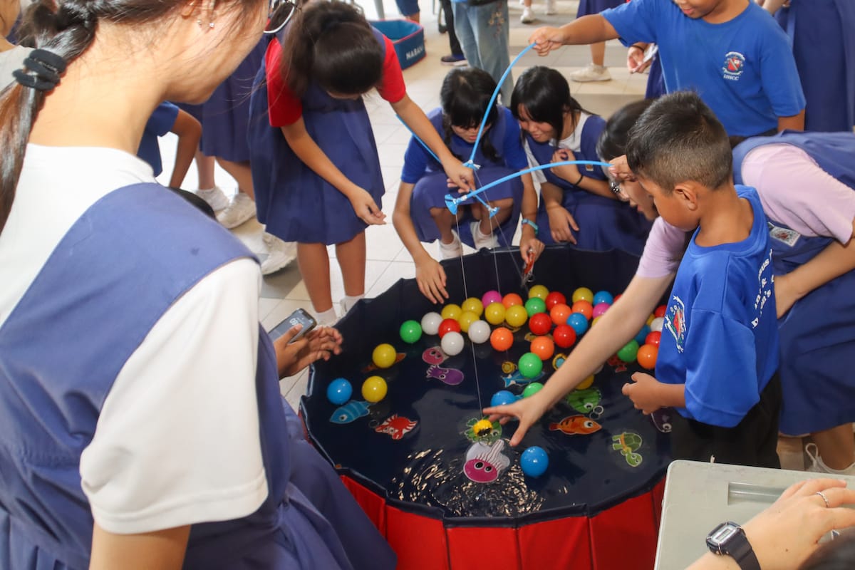 Children in blue uniforms gather around a small pool filled with colorful balls, playing a fishing game.